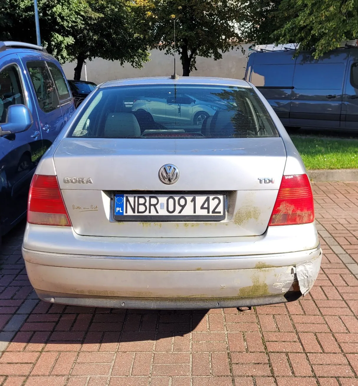 Tylny widok srebrnego samochodu Volkswagen Bora TDI zaparkowanego na kostce brukowej. Samochód ma polską tablicę rejestracyjną o numerze NBR 09142. Na tylnym zderzaku po prawej stronie widoczne jest uszkodzenie lakieru i odwarstwiająca się folia lub naklejka. Po lewej stronie znajduje się niebieski samochód, a w tle ciemnoszary furgon oraz zielone drzewa.
