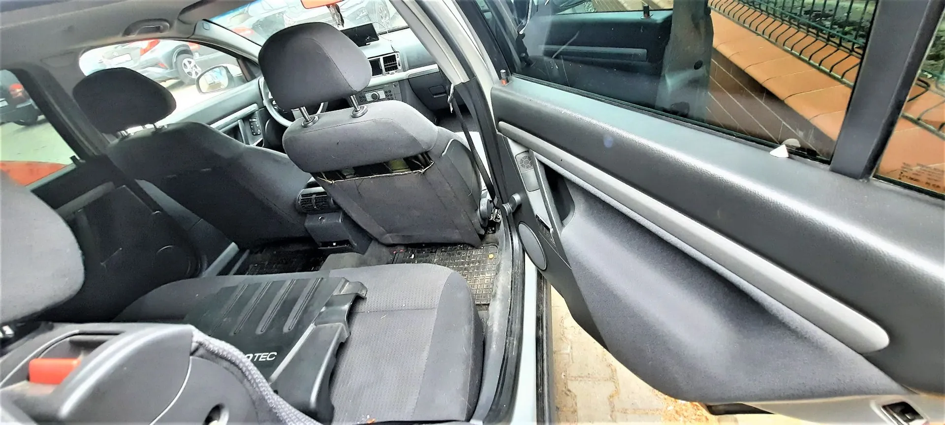 Wnętrze samochodu widziane z otwartych drzwi pasażera z przodu. Widoczne są ciemnoszare fotele materiałowe, w tym fotel kierowcy i pasażera oraz tylna kanapa. W dolnej części obrazu znajduje się fragment tylnego siedzenia z widocznym napisem 'OTEC'. W tle widać fragment deski rozdzielczej, kierownicę oraz widok przez przednią szybę na inne zaparkowane samochody.