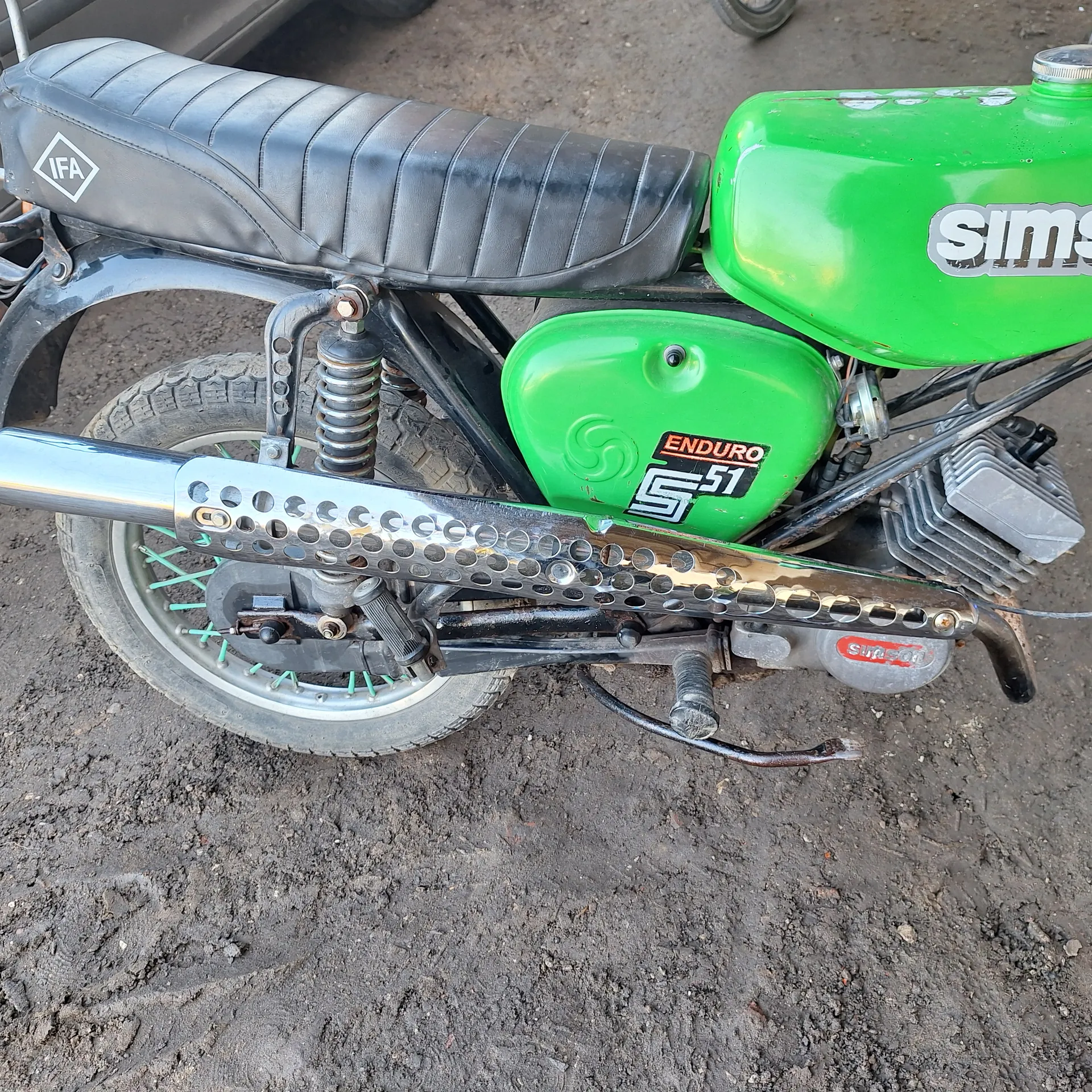 Zbliżenie na fragment motocykla z zielonymi zbiornikami paliwa, czarnym siedzeniem, metalowym wydechem z otworami i kołem tylnym z zielonymi promieniami. Tło stanowi brudna ziemia.