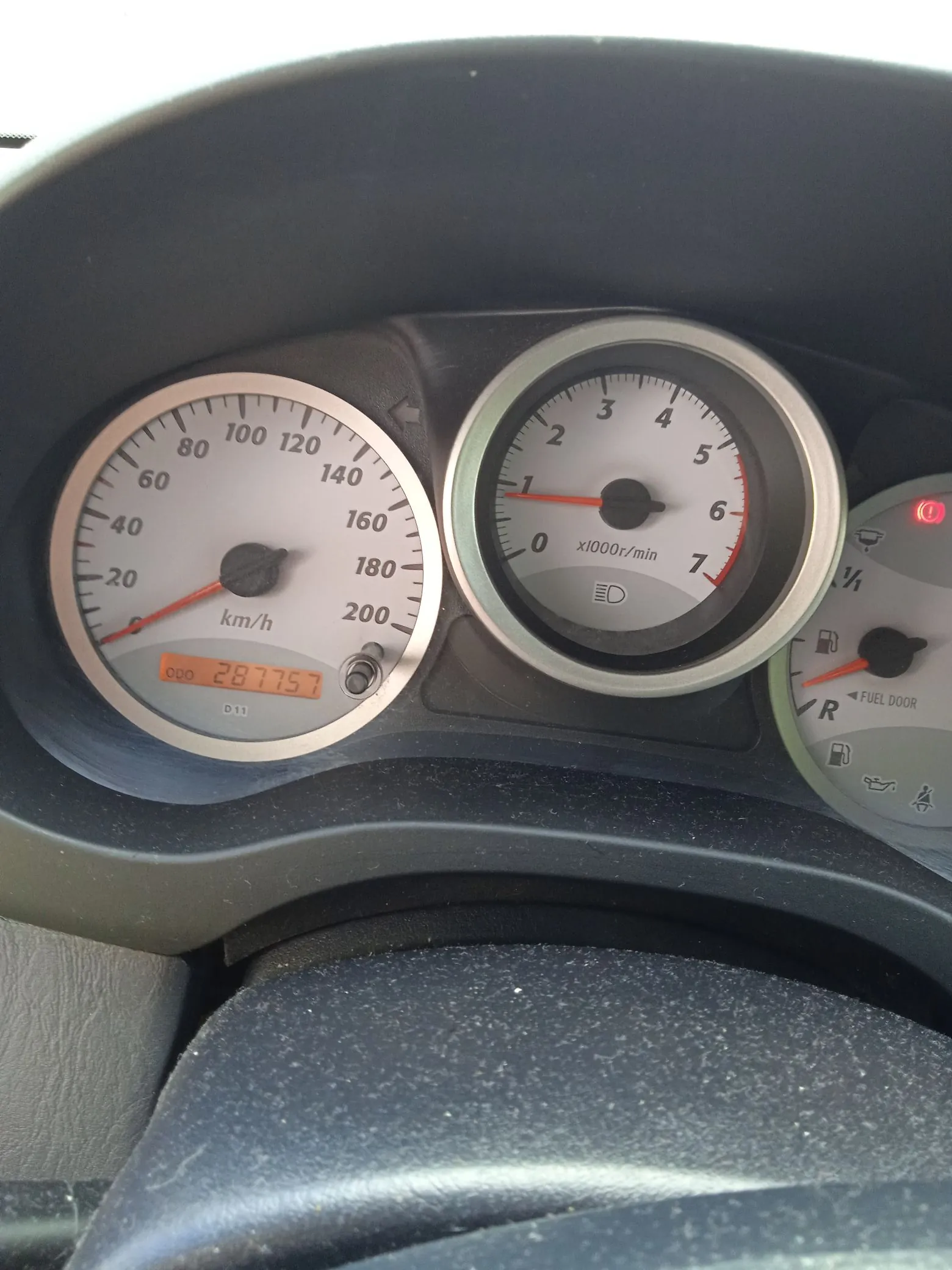 Zbliżenie na deskę rozdzielczą samochodu. Widoczny jest prędkościomierz z maksymalną wartością 200 km/h, wskazujący prędkość 0, oraz obrotomierz z maksymalną wartością 7 x1000r/min. Poniżej prędkościomierza znajduje się cyfrowy licznik przebiegu wyświetlający wartość 287757. Po prawej stronie widoczny jest wskaźnik poziomu paliwa z napisem FUEL DOOR oraz ikonami kontrolnymi.