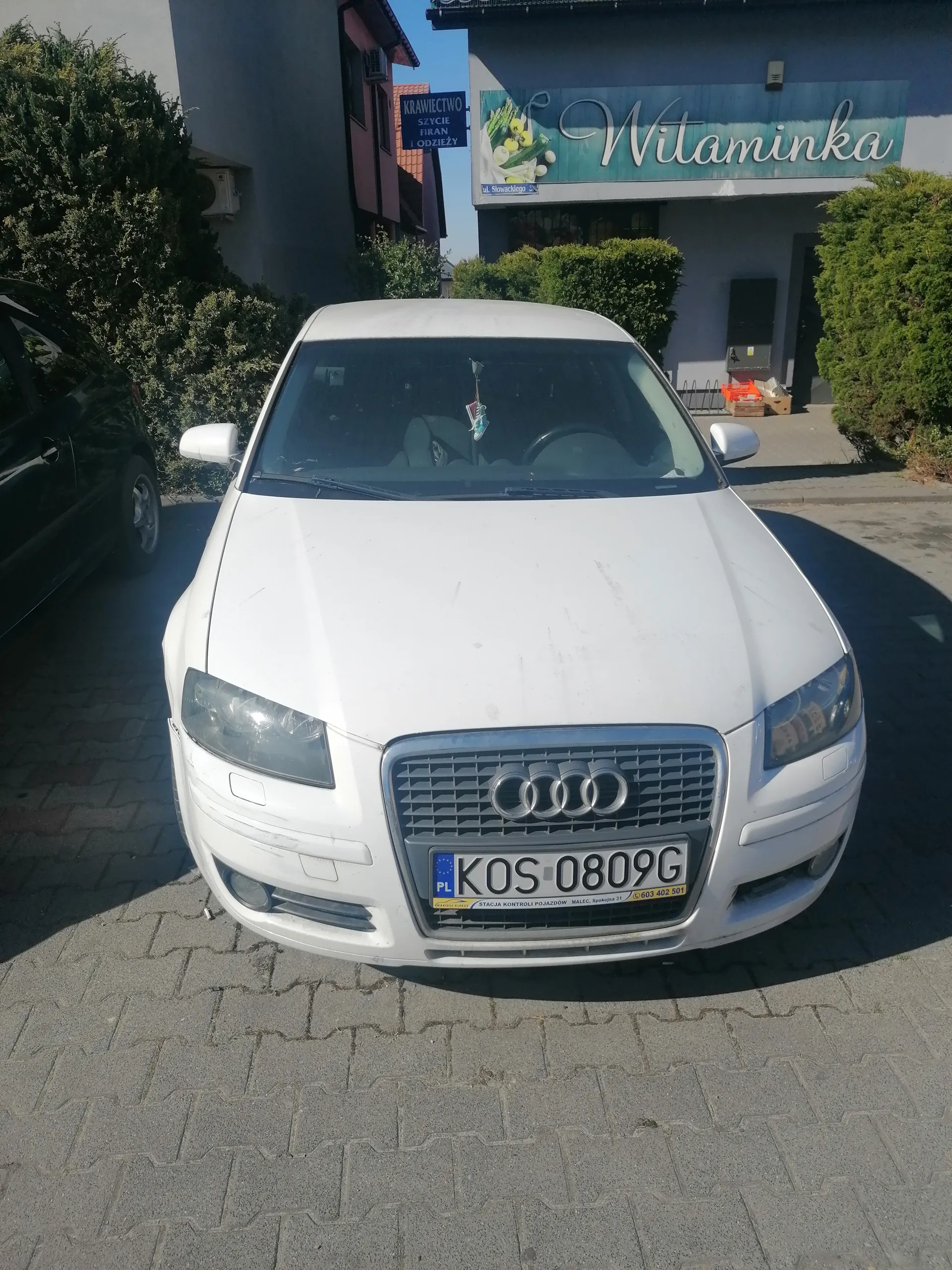 Biały samochód marki Audi z tablicą rejestracyjną KOS-0809G zaparkowany na brukowanej powierzchni. W tle budynek z niebieskim szyldem "Witaminka" oraz tablicą z napisem "Krawiectwo Szycie Firan Odzieży". Po lewej stronie częściowo widoczny czarny samochód. Wokół budynku krzewy i drzewa. Na tablicy rejestracyjnej widoczny napis "PL" oraz żółty napis na dole z numerem telefonu.