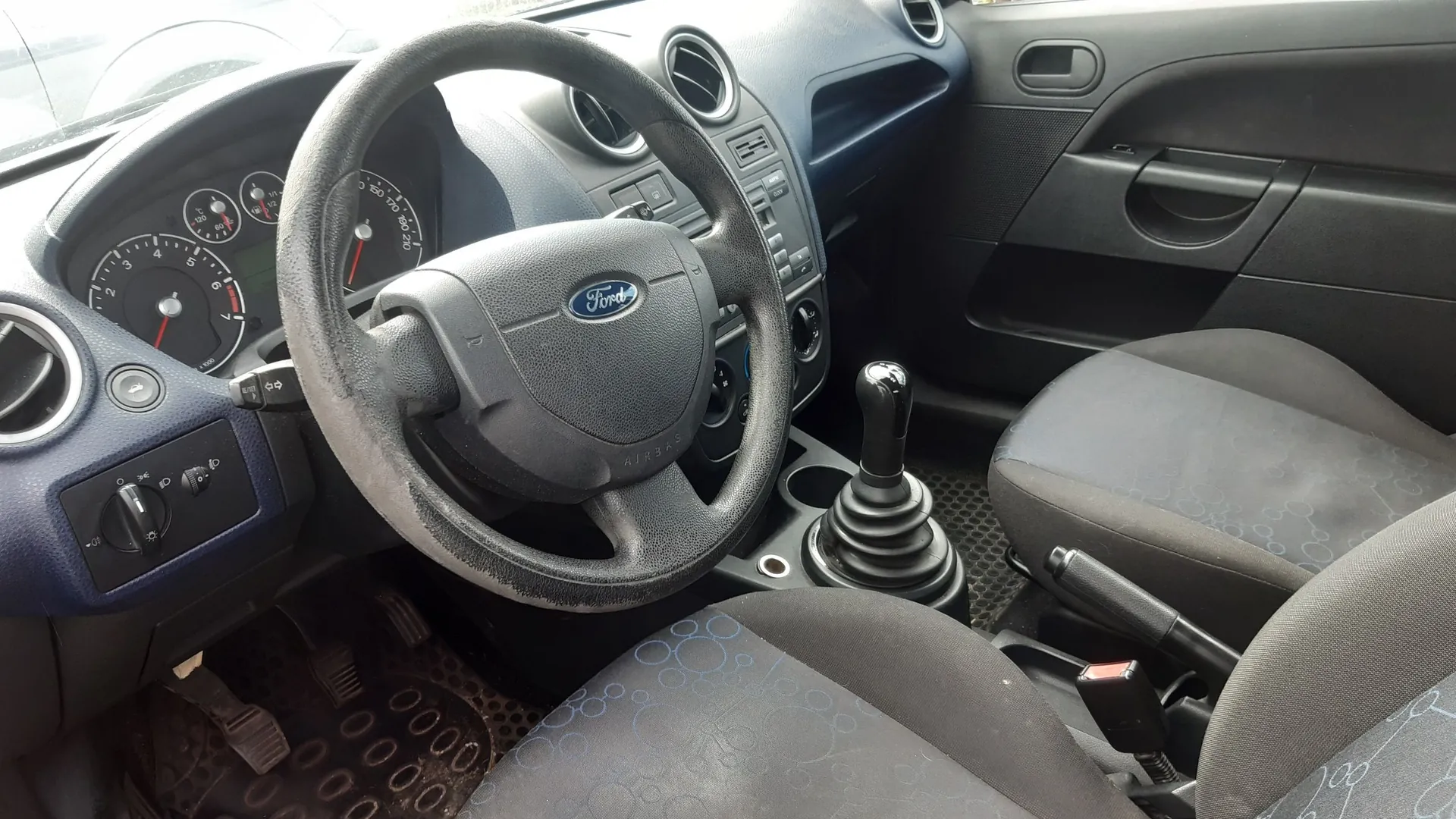 Wnętrze samochodu marki Ford z perspektywy kierowcy. Widoczna jest ciemnoszara kierownica z logo Ford, deska rozdzielcza z zegarami, centralna konsola z radiem i nawiewami oraz dwa przednie fotele z ciemną tapicerką w delikatny, niebieski wzór kółek. Widać również drążek zmiany biegów oraz hamulec ręczny.