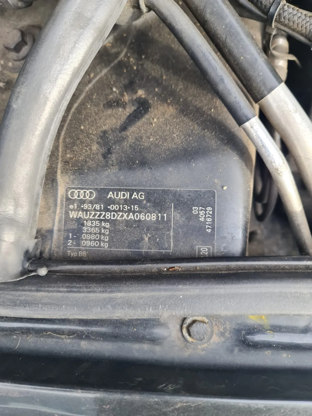Zbliżenie na tabliczkę identyfikacyjną samochodu marki Audi AG. Widoczne są cztery pierścienie logo Audi, napis 'AUDI AG', oznaczenia 'e1 93/81 0013 15', numer VIN 'WAUZZZ8DZXA060811', wartości '1835 kg', '3365 kg', '0980 kg', '0960 kg', oznaczenia '1-', '2-' oraz 'Typ B5'. Po prawej stronie tabliczki znajdują się dodatkowe numery: '03', '4057', '4716729' oraz kwadrat z napisem '20'. W tle widoczne są metalowe przewody i elementy konstrukcyjne. Tabliczka umieszczona jest na czarnym, lekko zabrudzonym powierzchni.