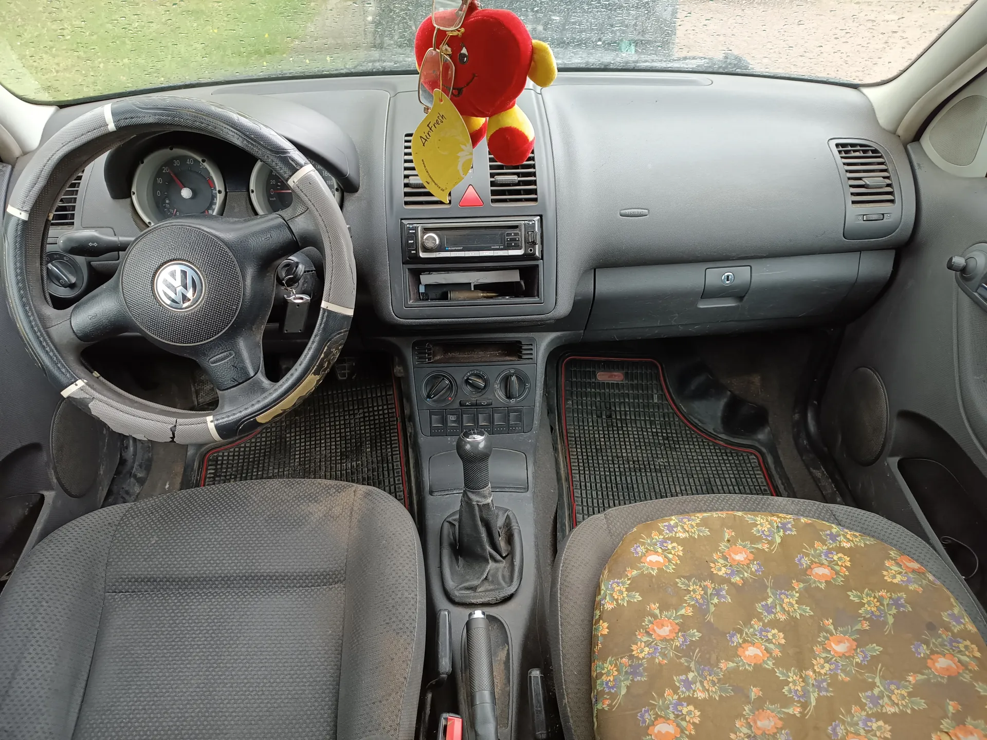 Wnętrze samochodu z widoczną kierownicą marki Volkswagen po lewej stronie, deska rozdzielcza z radioodtwarzaczem, wentylatorami i czerwonym pluszowym zabawkiem z żółtą tabliczką z napisem AirFresh wiszącym na lusterku. Przednie siedzenia: pasażerskie z kwiecistą pokrywką, kierowcy szare. W centrum deski rozdzielczej znajduje się dźwignia zmiany biegów, a na podłodze czarne dywaniki z czerwonym wykończeniem. Przez przednią szybę widoczny zielony teren i krople deszczu.