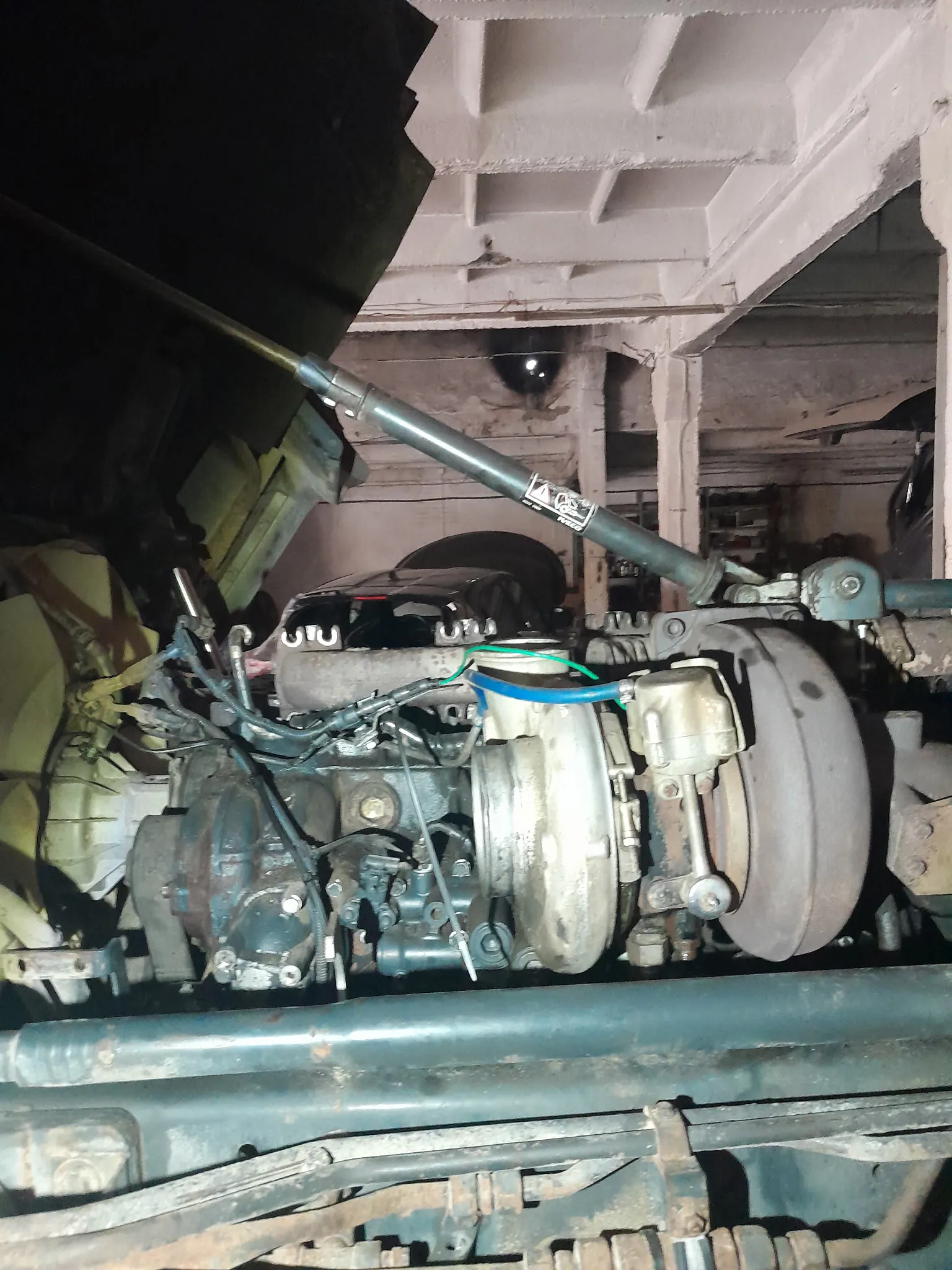 Zbliżenie na złożony układ mechaniczny, prawdopodobnie silnik lub podwozie pojazdu. Widoczne są metalowe rury, przewody (w tym niebieski i zielony), cylindryczne elementy oraz zardzewiałe części. Na jednej z rur znajduje się etykieta z nieczytelnym tekstem i symbolami. Tło stanowi warsztat z betonowymi ścianami i belkami sufitowymi. Zdjęcie wykonane z niskiej perspektywy, ukazujące dolną część konstrukcji.