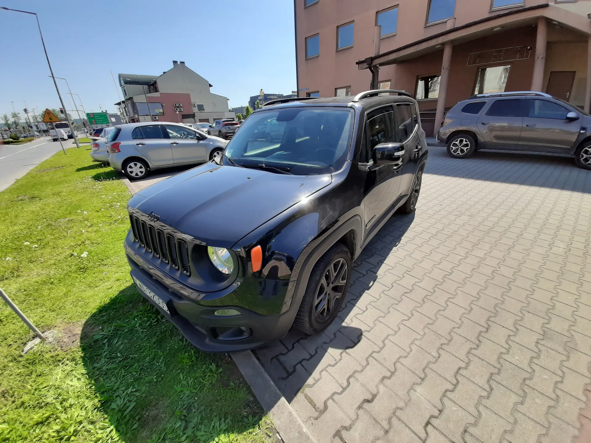 Czarny samochód Jeep zaparkowany na brukowanej powierzchni obok trawnika. W tle srebrny samochód osobowy, szary SUV, budynki, droga z sygnalizacją świetlną i znakami drogowymi. Jasne niebo bez chmur.