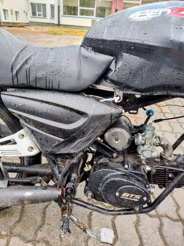 Zbliżenie na czarny motocykl z widocznymi kroplami wody na siedzeniu, zbiorniku paliwa i częściach mechanicznych. Na zbiorniku widoczny fragment napisu 'Ben', a na silniku napis 'BTS'. W tle budynek z oknami i brukowana powierzchnia. Motocykl stoi na chodniku z płytek w kształcie łuków.