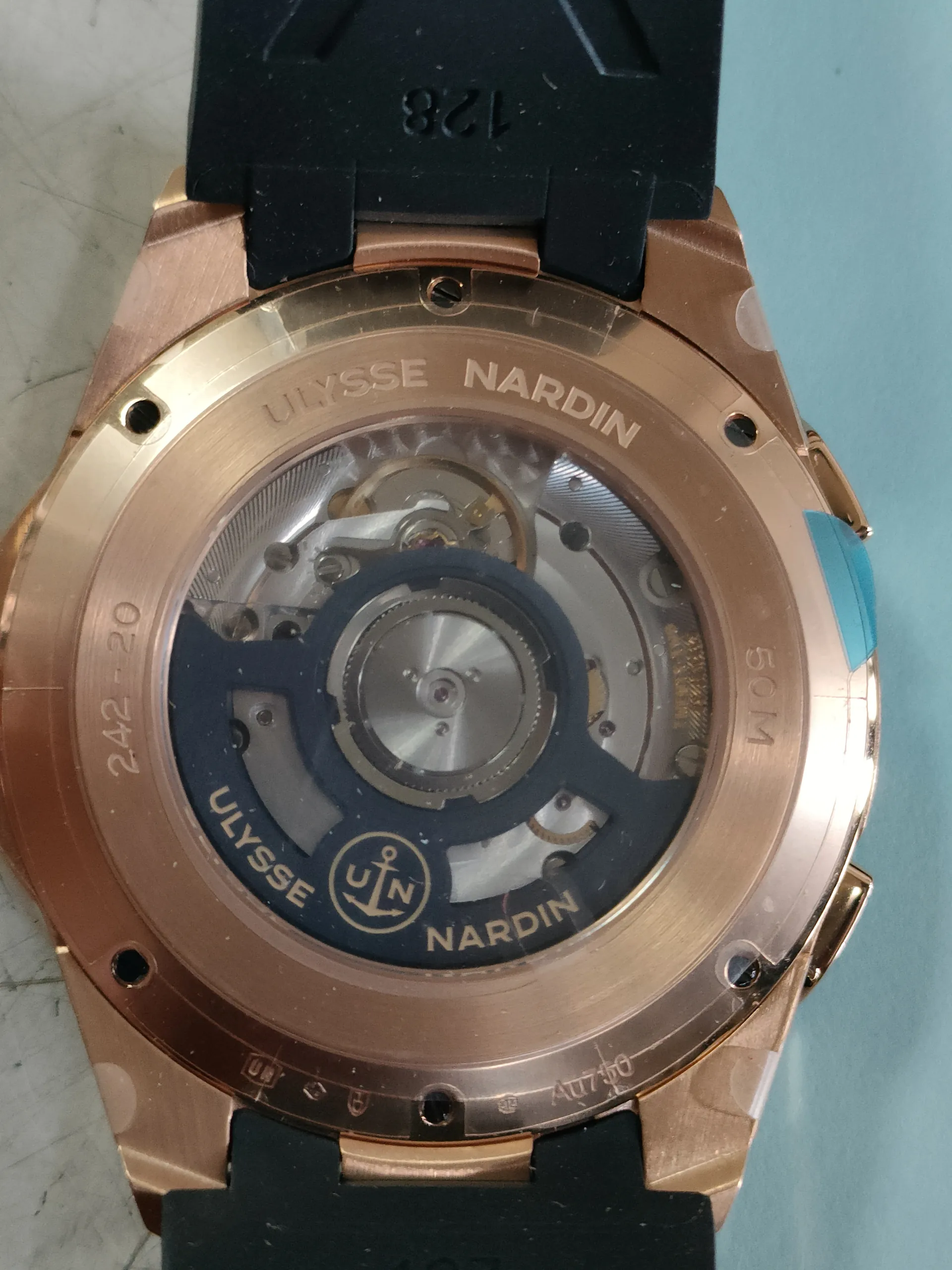 Zbliżenie na tylną część zegarka Ulysse Nardin z przezroczystą kopertą pokazującą mechanizm. Obudowa w kolorze różowego złota z widocznymi napisami: 'ULYSSE NARDIN' wzdłuż brzegu, '50M' u góry, '242-20' u dołu oraz logo z kotwicą i literami 'UN' w centrum. Czarny pasek z napisem '128' po lewej stronie. Tło w kolorze jasnoniebieskim.
