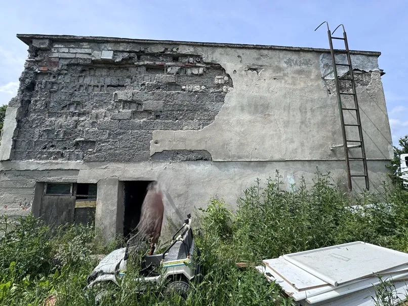 Zdjęcie przedstawia zniszczoną, betonową ścianę starego budynku z widocznymi ubytkami tynku i odsłoniętą strukturą z bloków. Po prawej stronie ściany przymocowana jest metalowa drabina prowadząca na dach. Przed budynkiem znajduje się gęsta, zielona roślinność, w której stoi biały pojazd typu quad oraz fragmenty białych płyt budowlanych leżące na ziemi.