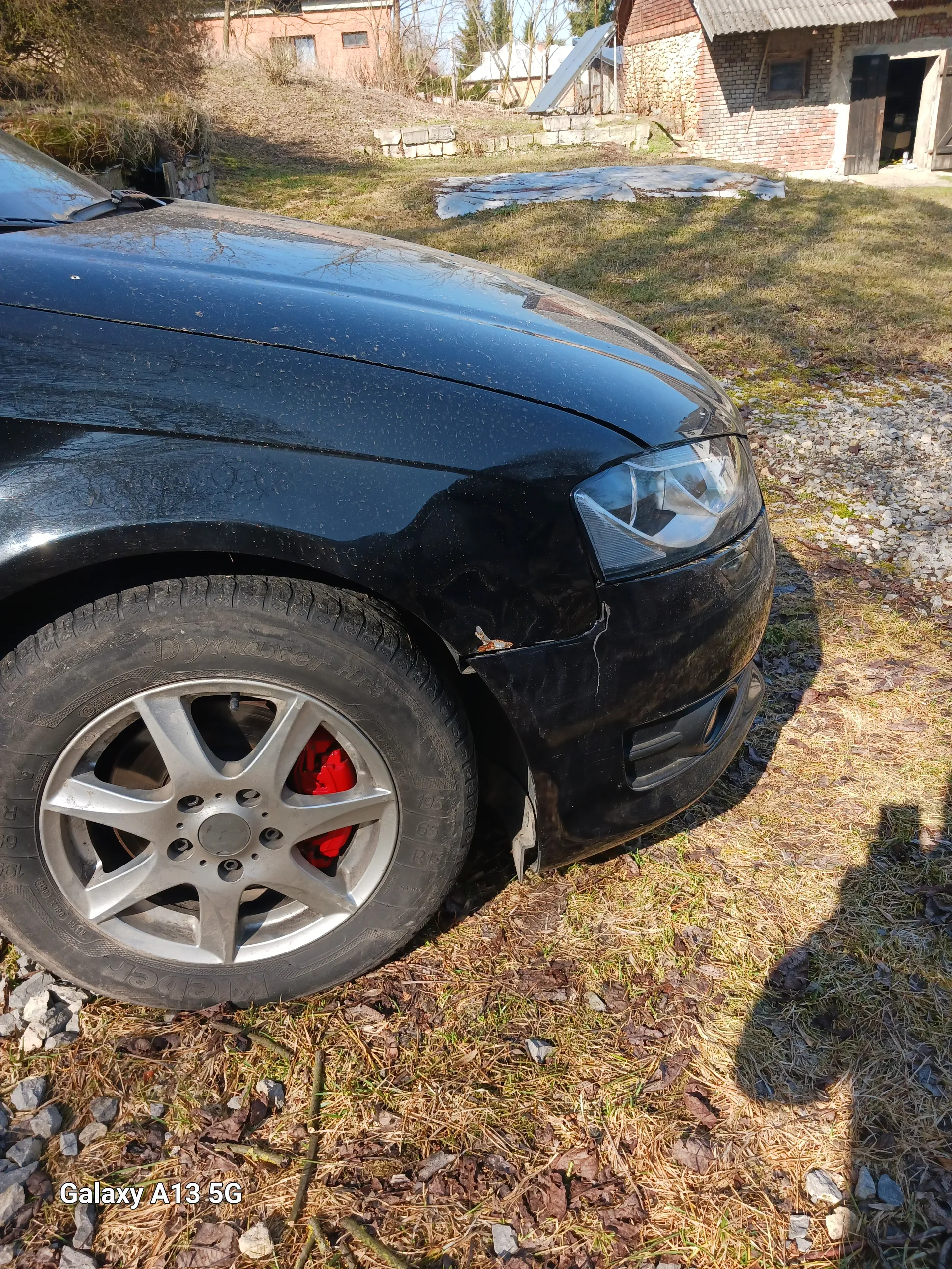 Zdjęcie przedstawia czarny samochód z widocznym przodem, w tym reflektorem i kołem z srebrnym felgiem oraz czerwonymi zaciskami hamulcowymi. Na przednim zderzaku widoczne są uszkodzenia. W tle dom z cegły, stos drewna, trawa i żwir. Po prawej stronie zdjęcia widoczny pionowy napis 'Galaxy A13 5G'.