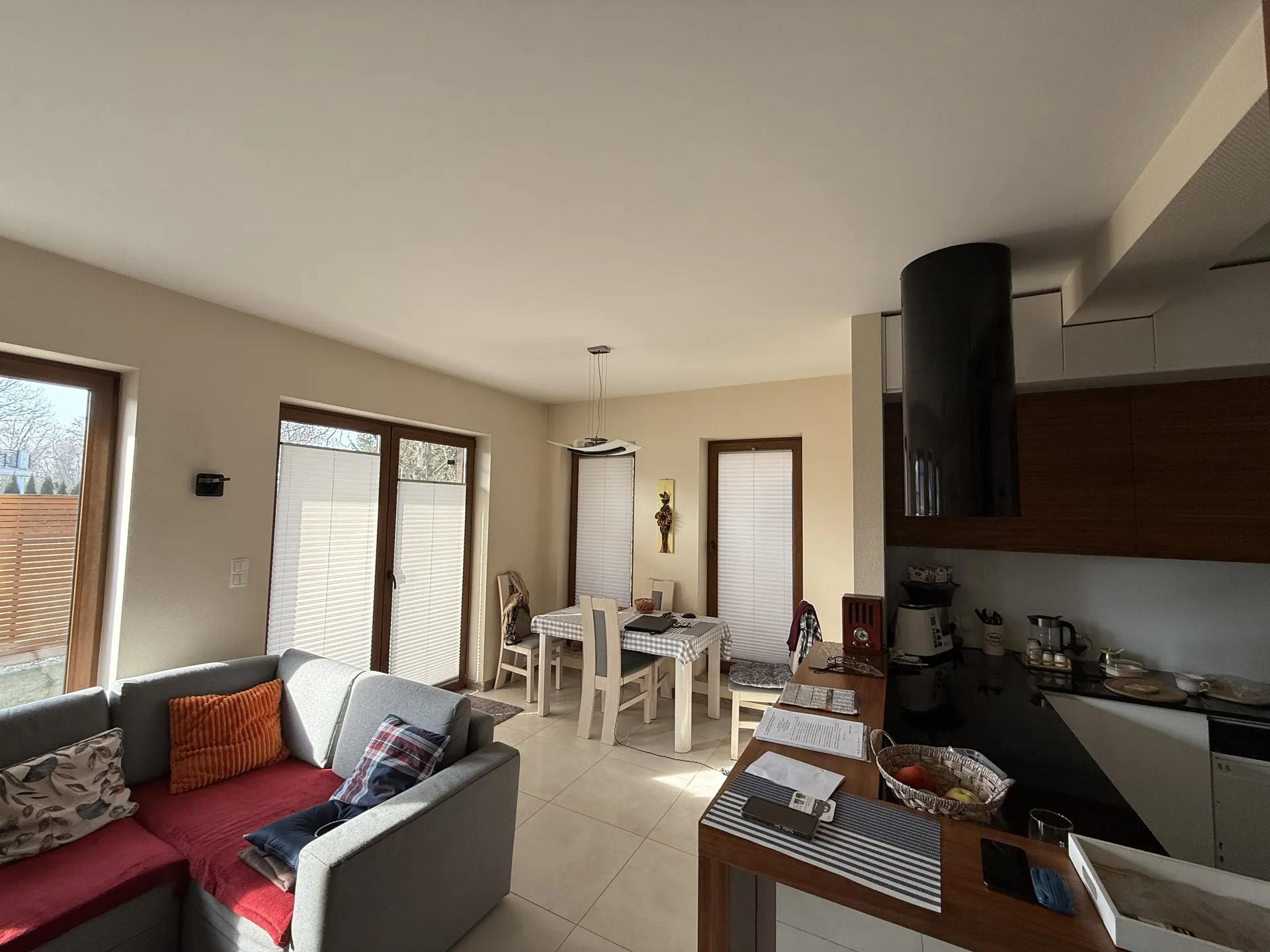 Wnętrze mieszkania z salonem i kuchnią. Po lewej stronie szara sofa z czerwonymi poduszkami i poduszkami w wzory. W tle stół z białymi krzesłami przykryty kratowaną serwetką. Okna z roletami, jedno otwarte na zewnątrz z widokiem na ogrodzenie. Po prawej stronie kuchnia z ciemnymi szafkami, czarną blatem i cylindrycznym kominem. Na blacie kuchennym: koszyk z owocami, czajnik, dokumenty, telefon i inne przedmioty. Podłoga z jasnych płytek, sufit biały. Ściany w kolorze beżu.