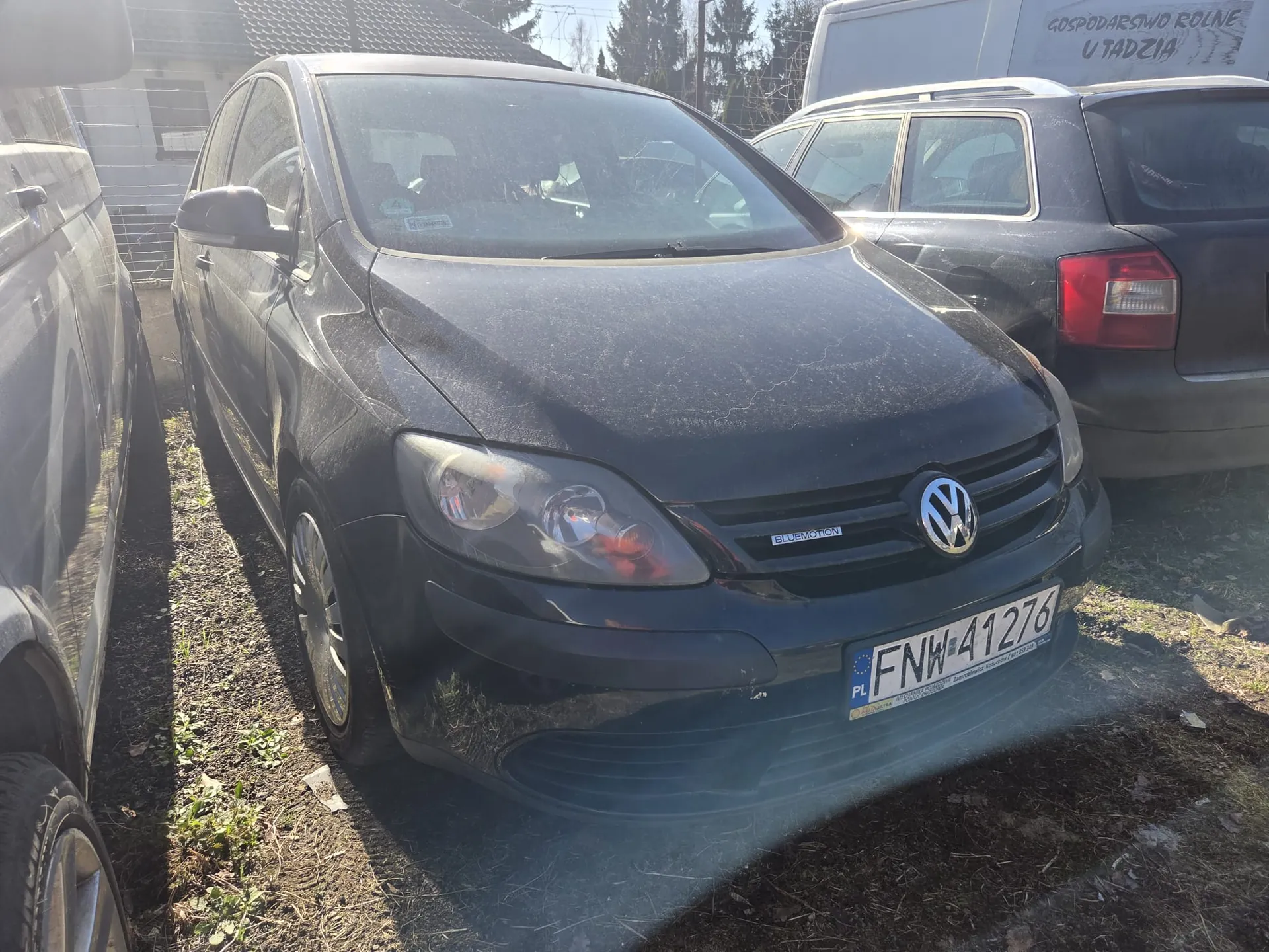 Czarny samochód marki Volkswagen z tablicą rejestracyjną PL FNW 41276 i napisem BLUEMOTION na masce. Pojazd stoi na podwórku z innymi samochodami: szarym po lewej i ciemnym po prawej. W tle budynek z napisem GOSPODARSTWO ROLNE U TADZIA oraz drzewa. Podłoże ziemne z fragmentami trawy. Samochód pokryty jest kurzem, widoczne reflektory i logo VW.