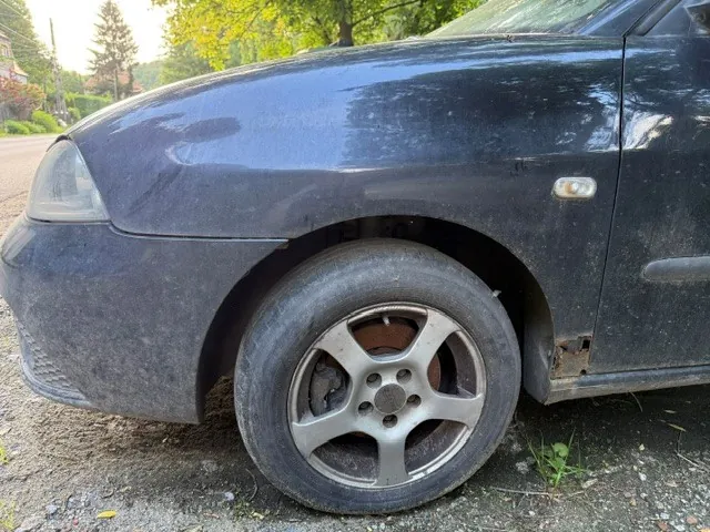 Zbliżenie na przednią lewą stronę czarnego samochodu. Widoczna jest przednia lampa, maska z widocznymi zarysowaniami oraz przednie koło z pięcioramienną srebrną felgą. W dolnej części nadkola, przy progu auta, widoczne jest uszkodzenie karoserii w postaci rdzy i ubytku blachy.
