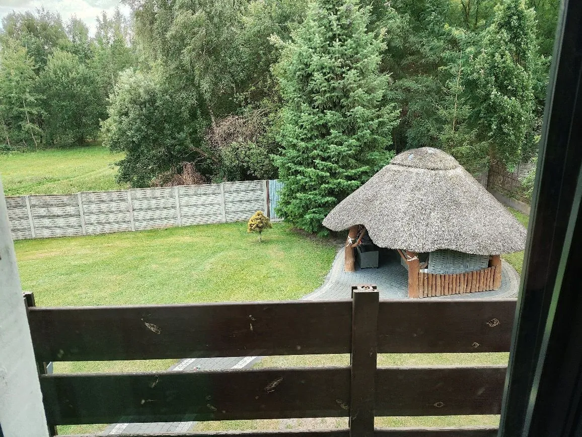Widok z balkonu na ogród z zieloną trawą. Po prawej stronie znajduje się altana z grubym, strzechowym dachem i drewnianym ogrodzeniem wokół podstawy. W tle widać betonowy płot oraz gęstą ścianę zielonych drzew i krzewów. Na pierwszym planie widoczna jest ciemnobrązowa, drewniana barierka balkonu.