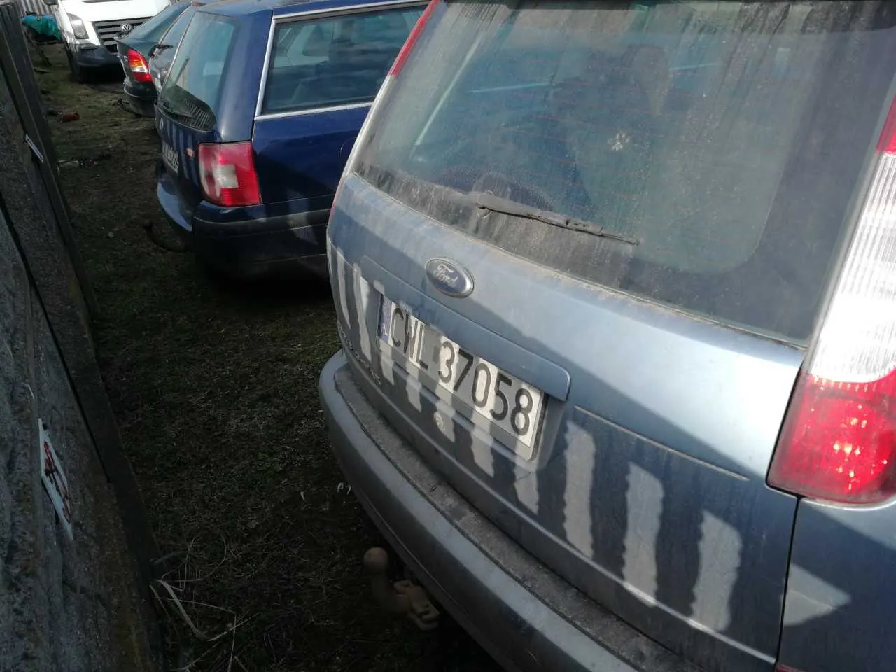 Zdjęcie przedstawia tył niebieskiego samochodu marki Ford z tablicą rejestracyjną CWL 37058. Po lewej stronie widoczna kamienna ściana z małym znakiem, a na tle kolejne zaparkowane pojazdy na trawie. Samochód znajduje się na trawiastym obszarze, a na jego tylnym szybie widoczne są ślady brudu.
