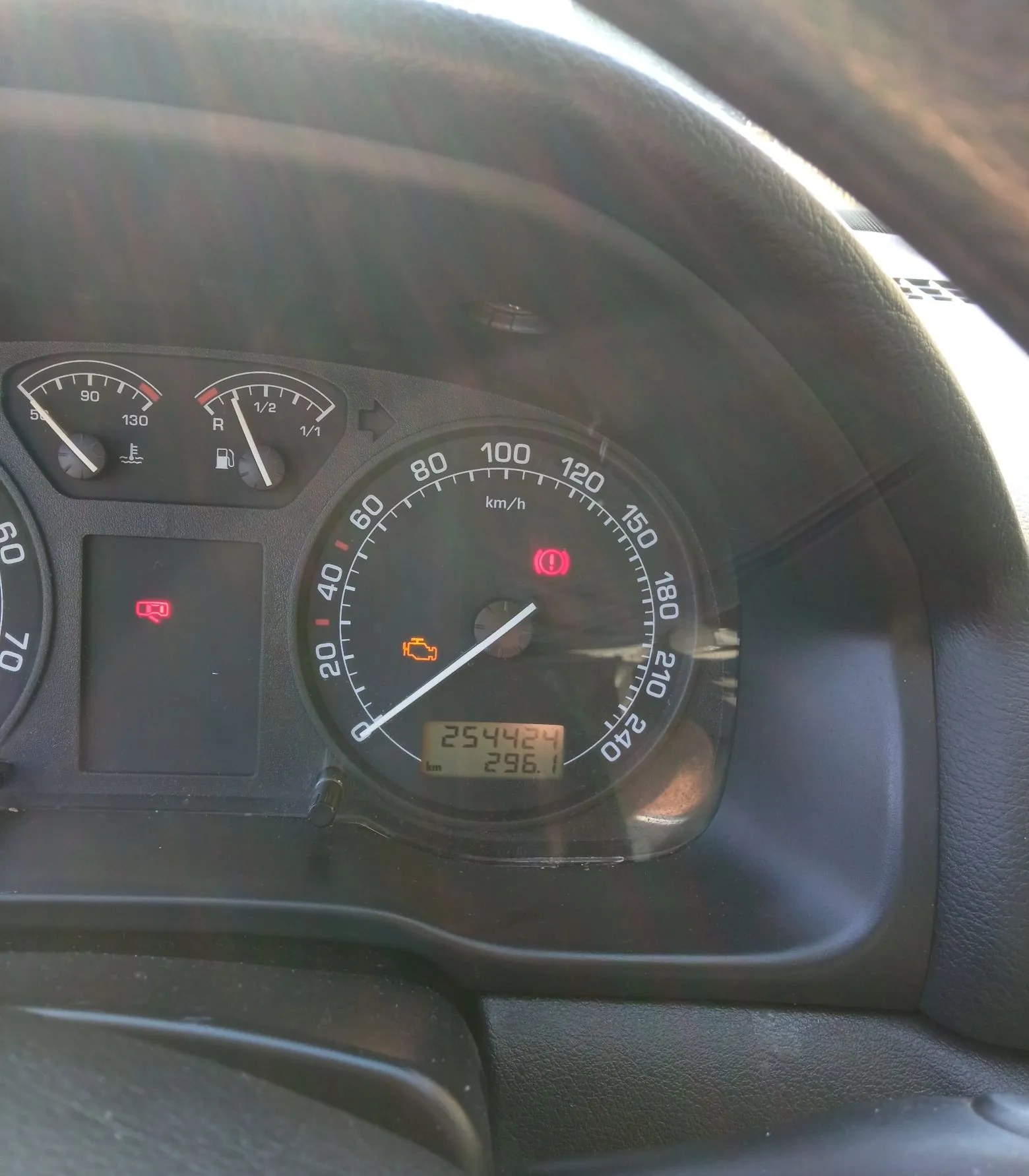 Zdjęcie z wnętrza samochodu przedstawiające panel instrumentów. Widoczny jest prędkościomierz z maksymalną wartością 240 km/h, wskazujący prędkość 0 km/h. Na tarczy prędkościomierza świecą się dwie czerwone kontrolki: ikona wykrzyknika w kółku oraz pomarańczowa kontrolka silnika. Cyfrowy licznik kilometrów wskazuje wartość 254424 km oraz 296.1 km. W tle widoczne są wskaźniki temperatury i poziomu paliwa.