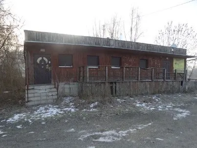 Jednokondygnacyjny, drewniany budynek o prostokątnym kształcie z płaskim dachem. Budynek posiada ciemne drzwi wejściowe po lewej stronie, kilka małych okien z ciemnymi żaluzjami oraz drewniany taras z barierką biegnący wzdłuż fasady. Po prawej stronie znajduje się żółta tablica informacyjna. Budynek stoi na fundamencie z betonowych bloczków, a przed nim znajduje się szutrowy plac z resztkami śniegu. W tle widoczne są bezlistne drzewa.