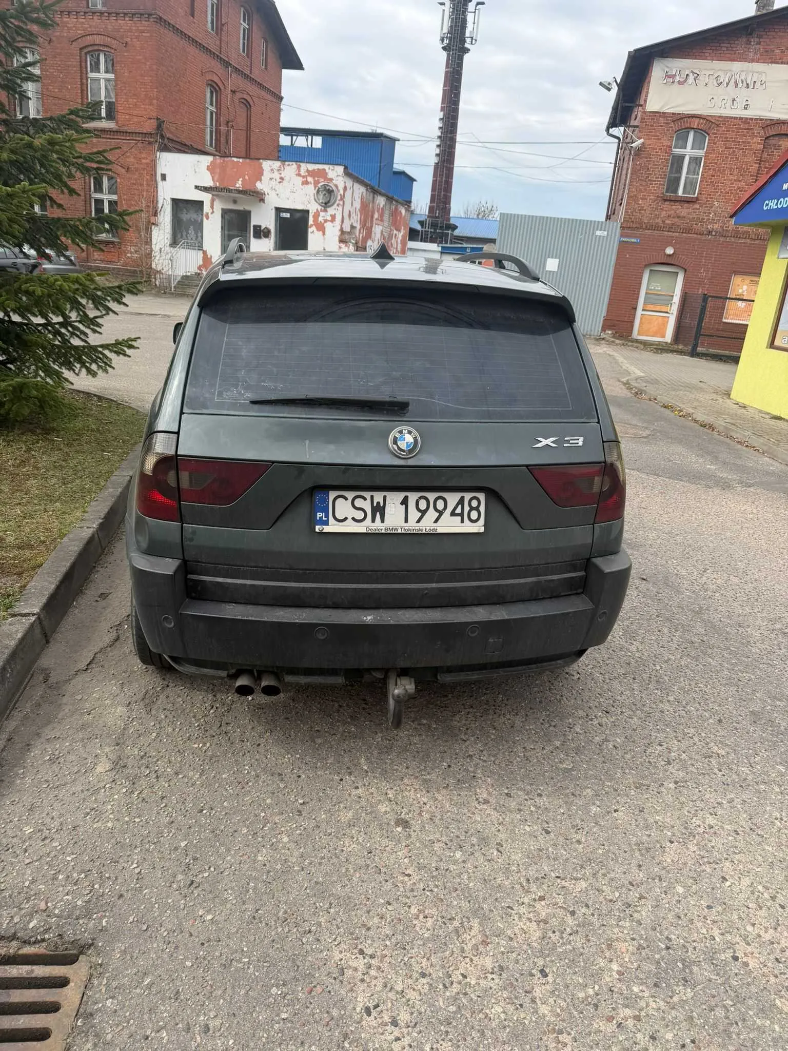 Z tyłu widoczny ciemnoszary samochód BMW X3 z tablicą rejestracyjną CSW 19948 (PL) oraz napisem "Dealer BMW Tłokinski Łódź" pod nią. Samochód stoi na asfaltowej drodze, po lewej stronie zielony krzew, w tle ceglane budynki, biały budynek z obłupioną farbą, wieża telekomunikacyjna oraz żółty budynek z niebieskim daszkiem. Niebo jest zachmurzone.