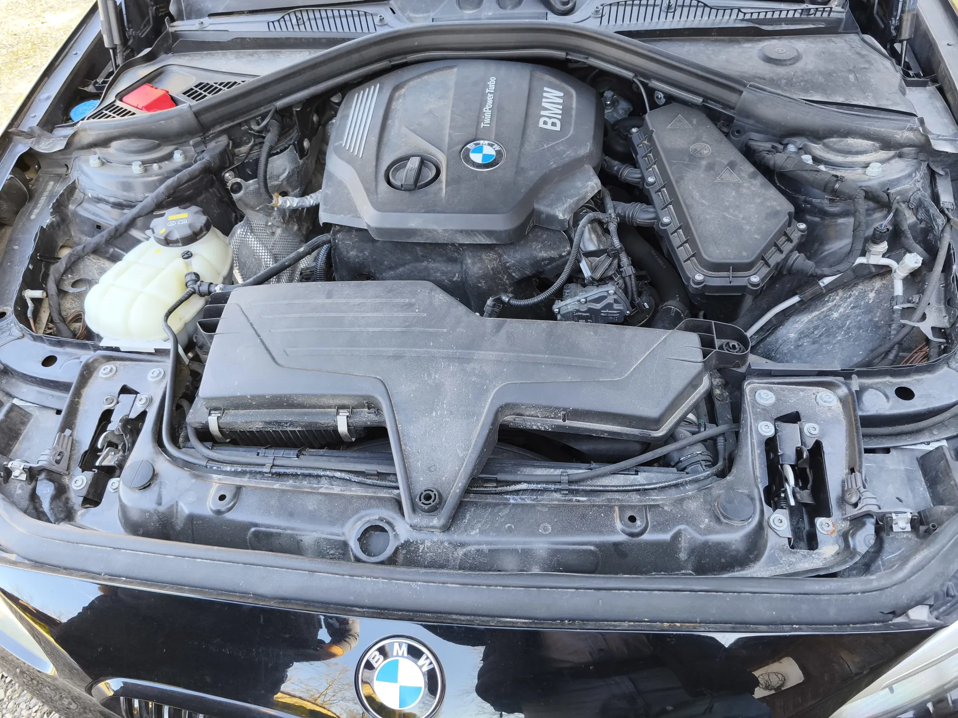 Otwarty kapota samochodu marki BMW, ukazujący komorę silnika. W centrum widoczna pokrywa silnika z napisem "BMW" i "TwinPower Turbo" oraz logo BMW. Po lewej stronie zbiornik chłodniczy z żółtawym płynem, czerwona pokrywka akumulatora oraz różne przewody i elementy mechaniczne. Na przedniej części pojazdu widoczne logo BMW na atrapie chłodnicy.