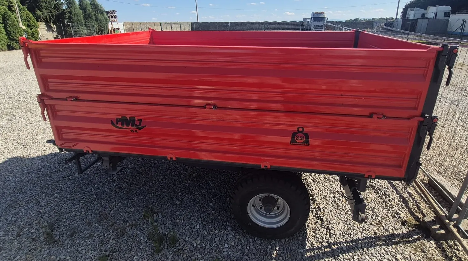 Czerwona przyczepa transportowa z metalową skrzynią o żebrowanych ściankach, stojąca na żwirowym podłożu. Na boku przyczepy widnieją napisy: logo 'PMJ' oraz tabliczka z napisem '3,5t ŁADOWNOŚĆ'. Przyczepa posiada jedno widoczne koło z szeroką oponą i srebrną felgą. W tle widać ogrodzenie, drzewa oraz zaparkowane ciężarówki pod jasnym, niebieskim niebem.
