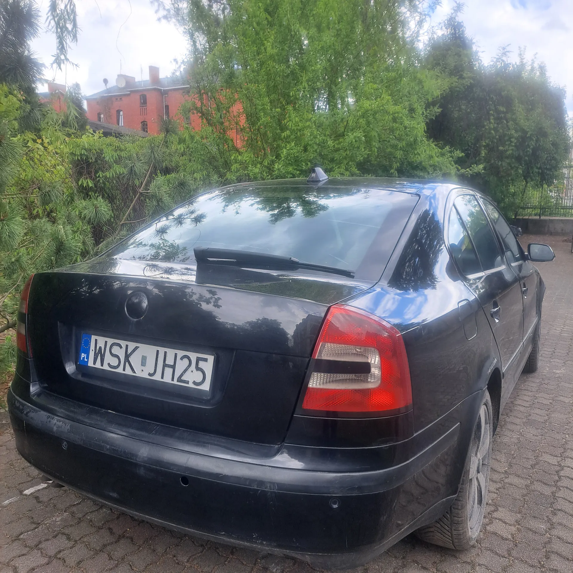 Czarny samochód widziany od tyłu z tablicą rejestracyjną WSK JH25, zaparkowany na brukowanej powierzchni. W tle zielone drzewa, czerwony budynek i częściowo zachmurzone niebo.