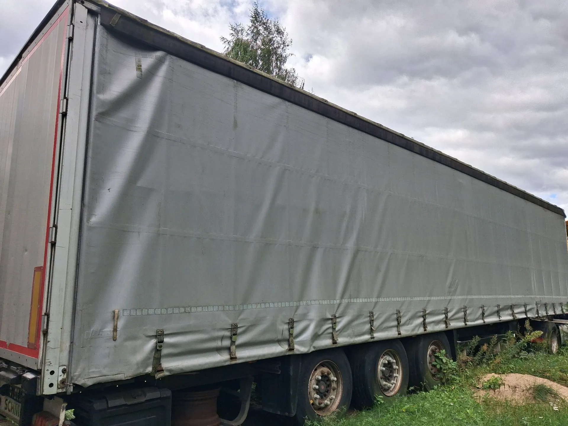 Boczny widok szarej naczepy ciężarówki z plandeką. Plandeka jest pognieciona w przedniej części i przymocowana do dołu za pomocą pasów z klamrami. Widoczne są trzy osie z kołami oraz fragmenty podwozia. W tle widać zieloną trawę, drzewo oraz zachmurzone niebo.