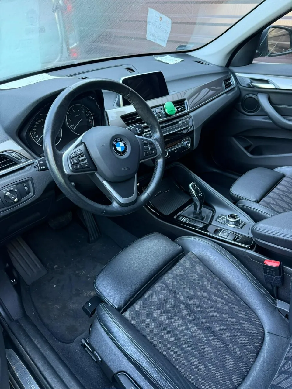 Wnętrze samochodu marki BMW z widocznym kierownicą z logo BMW, czarnymi skórzonymi fotelami z geometrycznym wzorem, deska rozdzielcza z licznikami, ekranem centralnym, przyciskiem start/stop w kolorze zielonym oraz dźwignią zmiany biegów. Na desce rozdzielczej i przedniej szybie znajdują się dokumenty. Podłoga w okolicy pedałów jest zabrudzona.