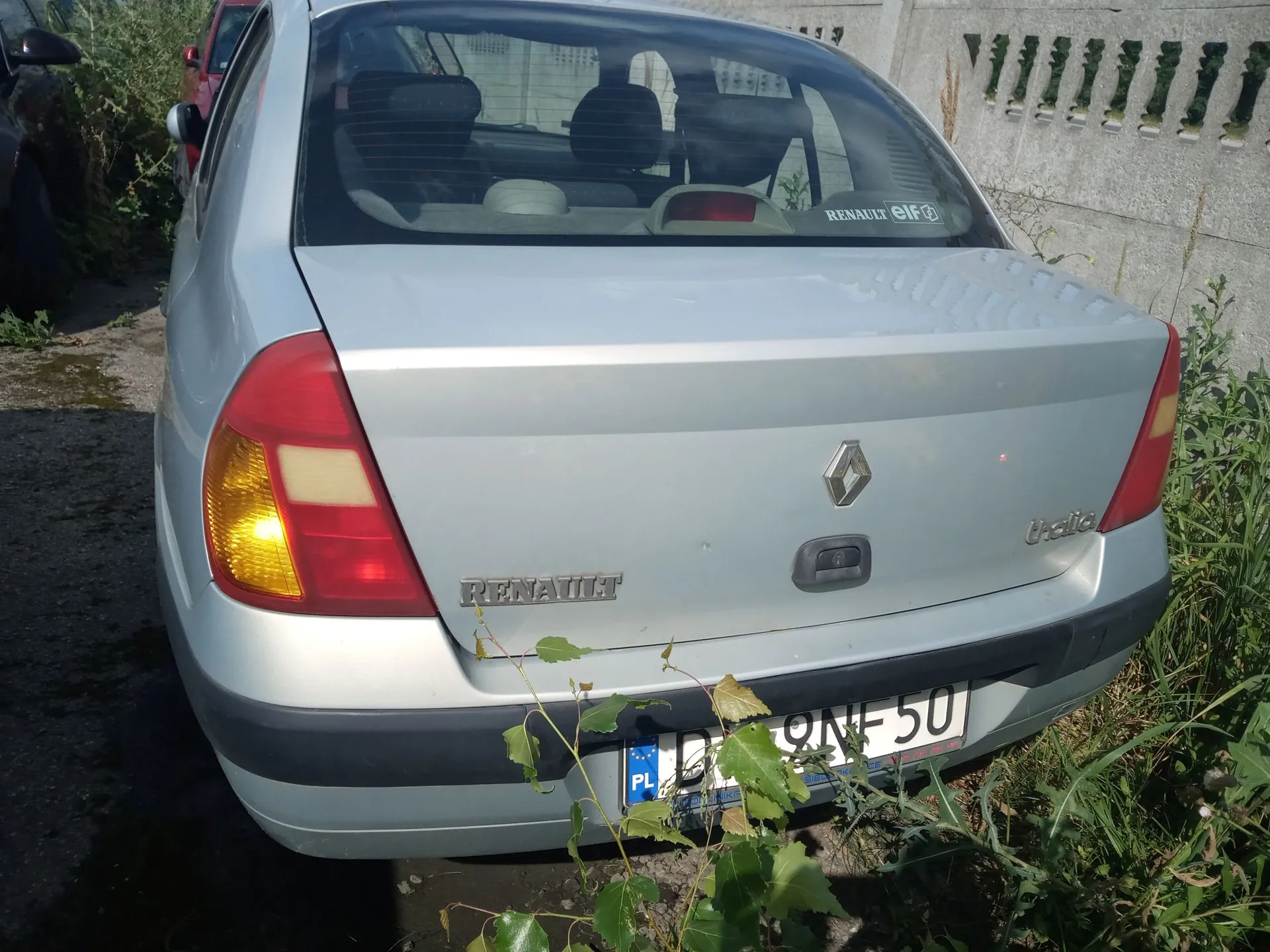 Tylny widok srebrnego samochodu marki Renault Symbol. Na klapie bagażnika widoczne jest logo Renault oraz napisy 'RENAULT' po lewej i 'Symbol' po prawej stronie. Samochód ma polską tablicę rejestracyjną z oznaczeniem 'PL', z której widoczne są znaki '9NF50'. Pojazd stoi zaparkowany obok zielonej roślinności i betonowego ogrodzenia.