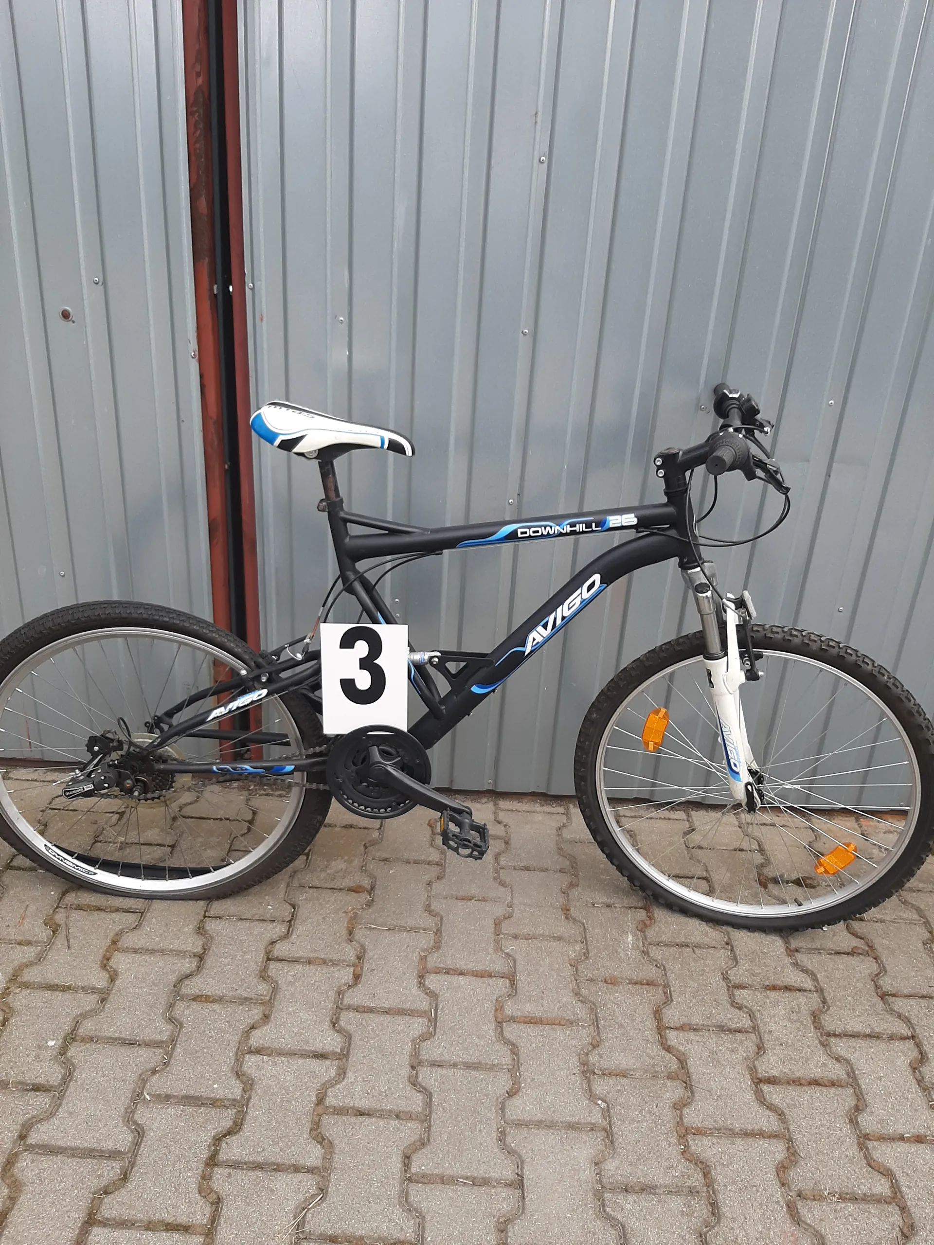 Czarny rower z niebieskimi akcentami marki AVIGO, model DOWNHILL 26, z białą tabliczką z czarną cyfrą 3. Rower umieszczony poziomo, oparty o szarą metalową ścianę i brukowaną nawierzchnię. Widoczne są pomarańczowe odblaski na przednim kole oraz biało-niebieskie siedzisko.