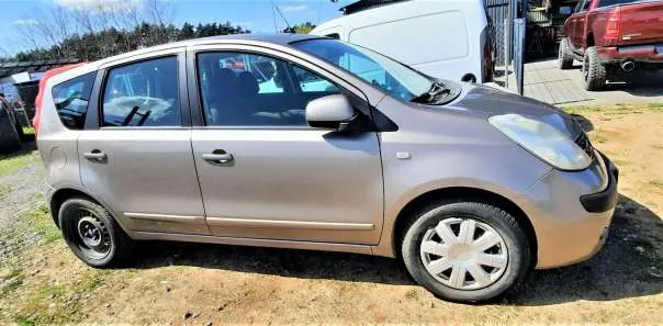 Srebrny samochód osobowy typu hatchback zaparkowany na terenie z żwirem i trawą. W tle widoczne są inne pojazdy: biały van i czerwony pickup, a także drzewa i budynki. Samochód ma czarne felgi, przednie światła i lusterka boczne. Niebo jest jasne, co wskazuje na słoneczną pogodę.