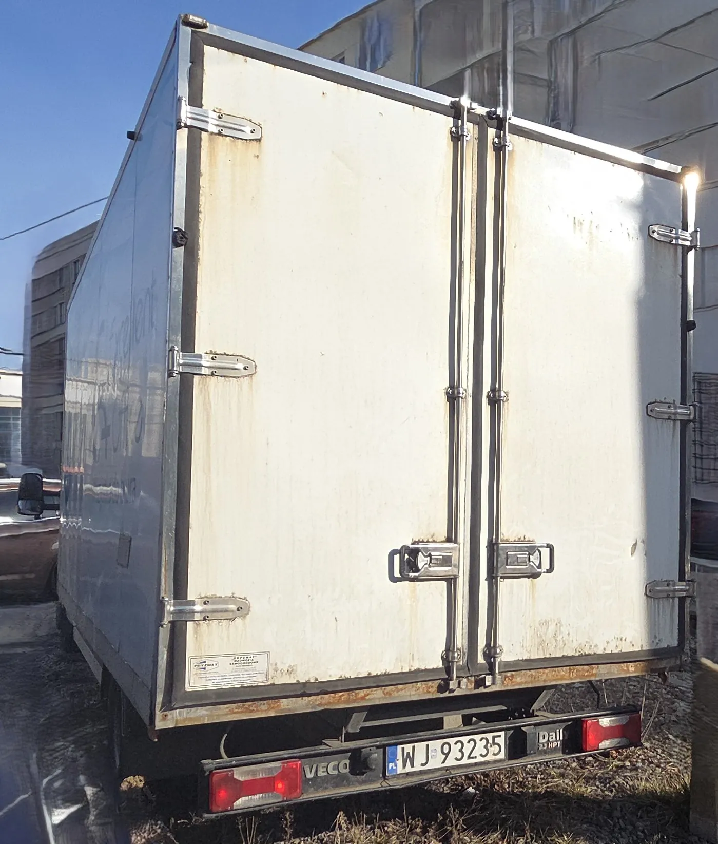 Tył białej ciężarówki z widocznymi metalowymi zawiasami i zatrzaskami. Na tylnej ścianie widoczne ślady rdzy, a na dole tablica rejestracyjna PL WJ 93235. Po lewej stronie napis VECO, po prawej Dall. W tle budynki i niebieskie niebo. Podwozie i tylne światła są czerwone.