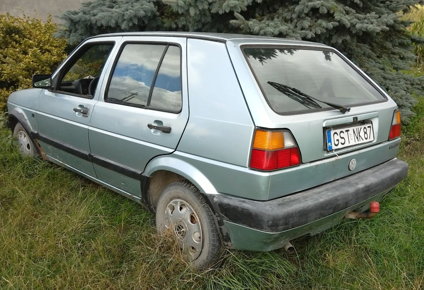 Srebrzysto-niebieski samochód Volkswagen Golf drugiej generacji zaparkowany na trawie przed zielonymi krzewami. Widoczny jest tył i bok auta z polską tablicą rejestracyjną o numerze GST NK 87. Samochód posiada czarne plastikowe zderzaki i listwy boczne.