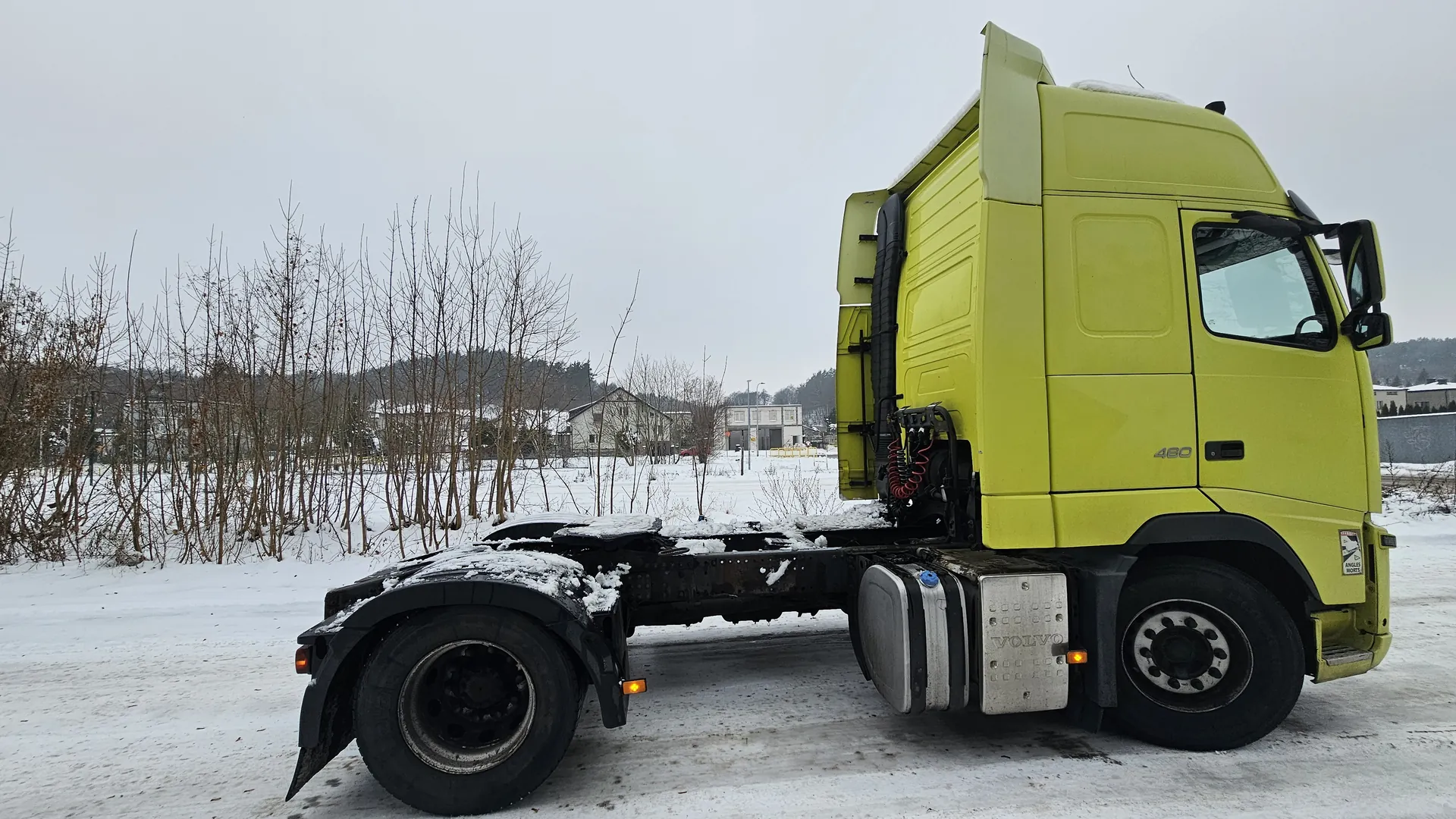 Żółta ciężarówka Volvo 460 bez przyczepy zaparkowana na śnieżnej drodze. Na nadwoziu, podwoziu i oponach widoczny śnieg. W tle nagie drzewa, domy, budynki oraz pochmurne niebo. Na boku pojazdu napisy "460" i "VOLVO".
