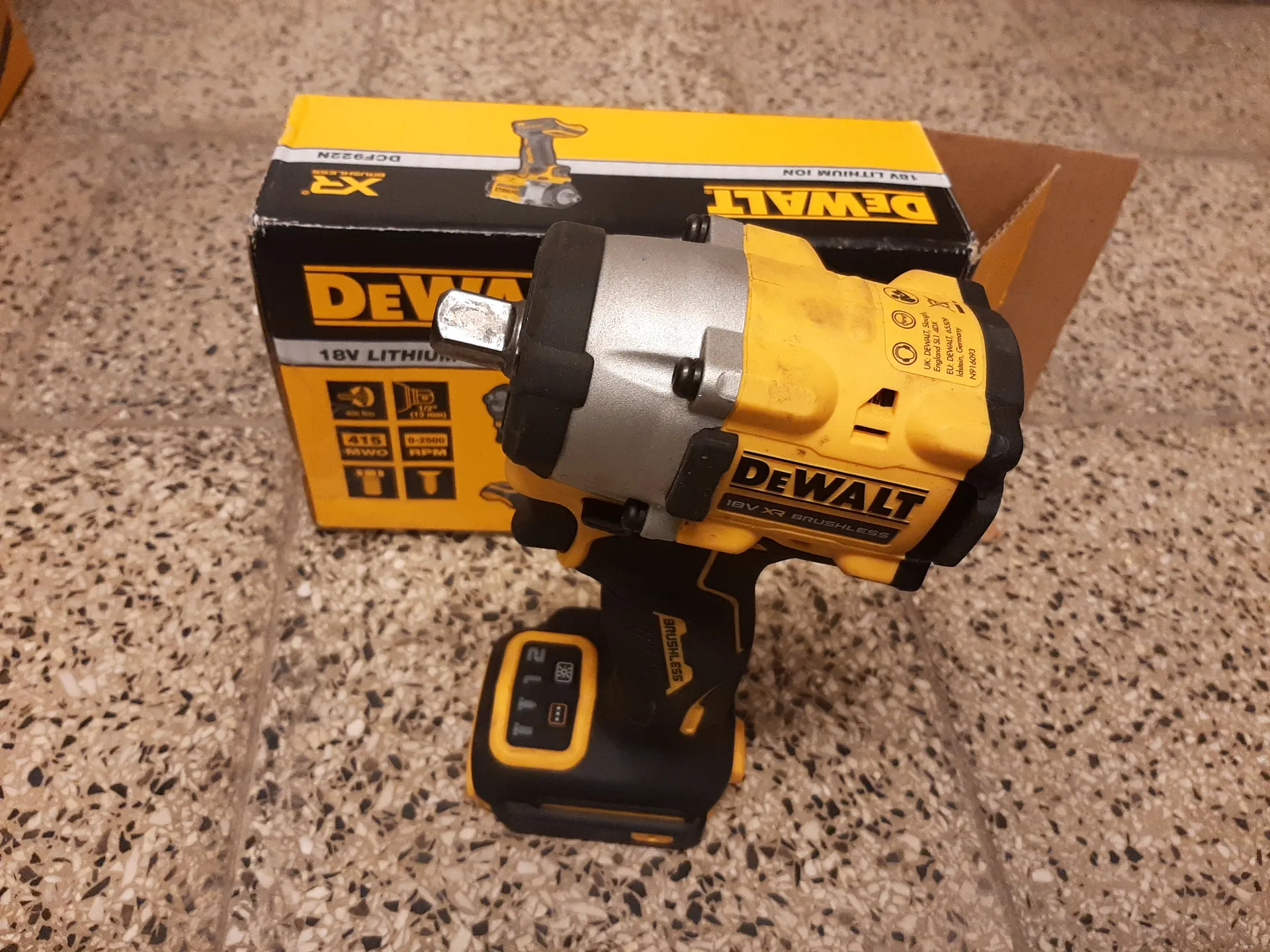 Żółto-czarny akumulatorowy wkrętarko-wkrętarka DeWALT 18V XR Brushless z akumulatorem pokazującym na wyświetlaczu cyfrę 21. W tle pudełko produktu z napisami DEWALT, 18V LITHIUM, XR oraz ikonami technicznymi. Całość umieszczona na podłodze z kafelkami w kolorze beżowym z czarnymi i białymi kropkami.