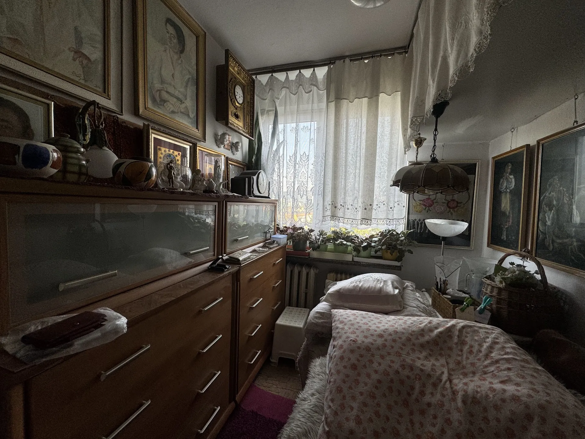 Pokój sypialny z łóżkiem przykrytym narzutą w kwiatowy wzór, poduszkami i białą pościelą. Po lewej stronie drewniane szafki z szklanymi frontami, na których umieszczono rama z obrazami, wazy oraz różne dekoracyjne przedmioty. Na ścianie wiszą liczne obrazy w ramach oraz zegar. Okno z koronkowymi zasłonami, na parapecie rozmieszczone doniczki z roślinami. W pokoju widoczna lampa wisząca oraz kosze z przedmiotami. Podłoga częściowo wyłożona dywanem.