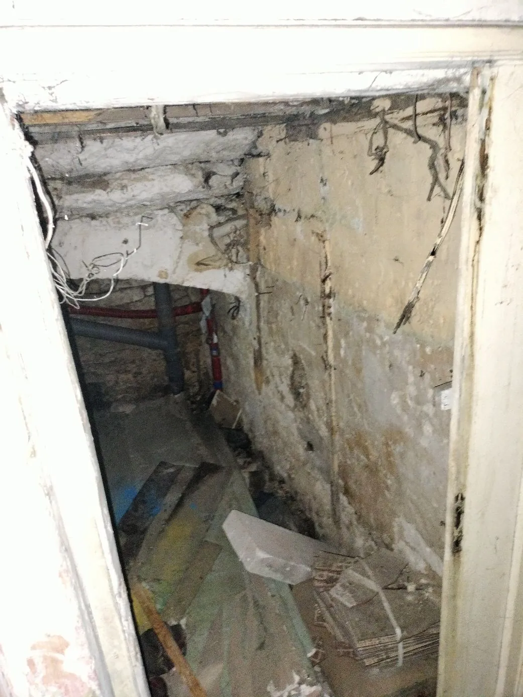Widok przez otwór drzwiowy do zniszczonego, surowego pomieszczenia przypominającego piwnicę lub schowek. Ściany i sufit są z betonu i tynku z licznymi ubytkami, zaciekami i odpadającym materiałem. W głębi widoczne są rury instalacyjne, a na podłodze leżą gruzy, kawałki płyt gipsowo-kartonowych i inne odpady budowlane.