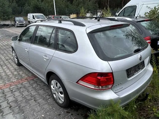Srebrny samochód osobowy marki Volkswagen Golf w wersji kombi, zaparkowany na kostce brukowej. Pojazd jest widoczny od tyłu i z boku. W tle znajdują się inne samochody, w tym biały furgon, oraz pas zieleni z drzewami.