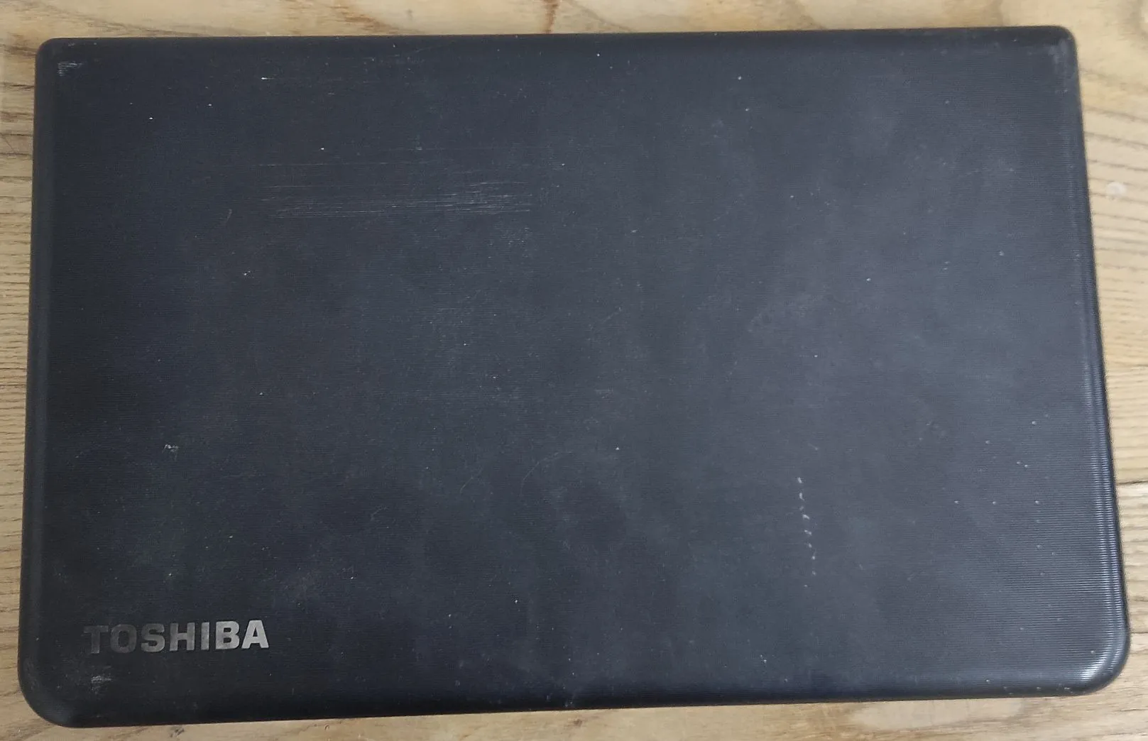 Widok z góry na zamkniętą pokrywę czarnego laptopa marki Toshiba. W lewym dolnym rogu znajduje się srebrny napis 'TOSHIBA'. Obudowa jest matowa i posiada widoczne ślady użytkowania w postaci drobnych zarysowań. Laptop leży na drewnianej powierzchni.
