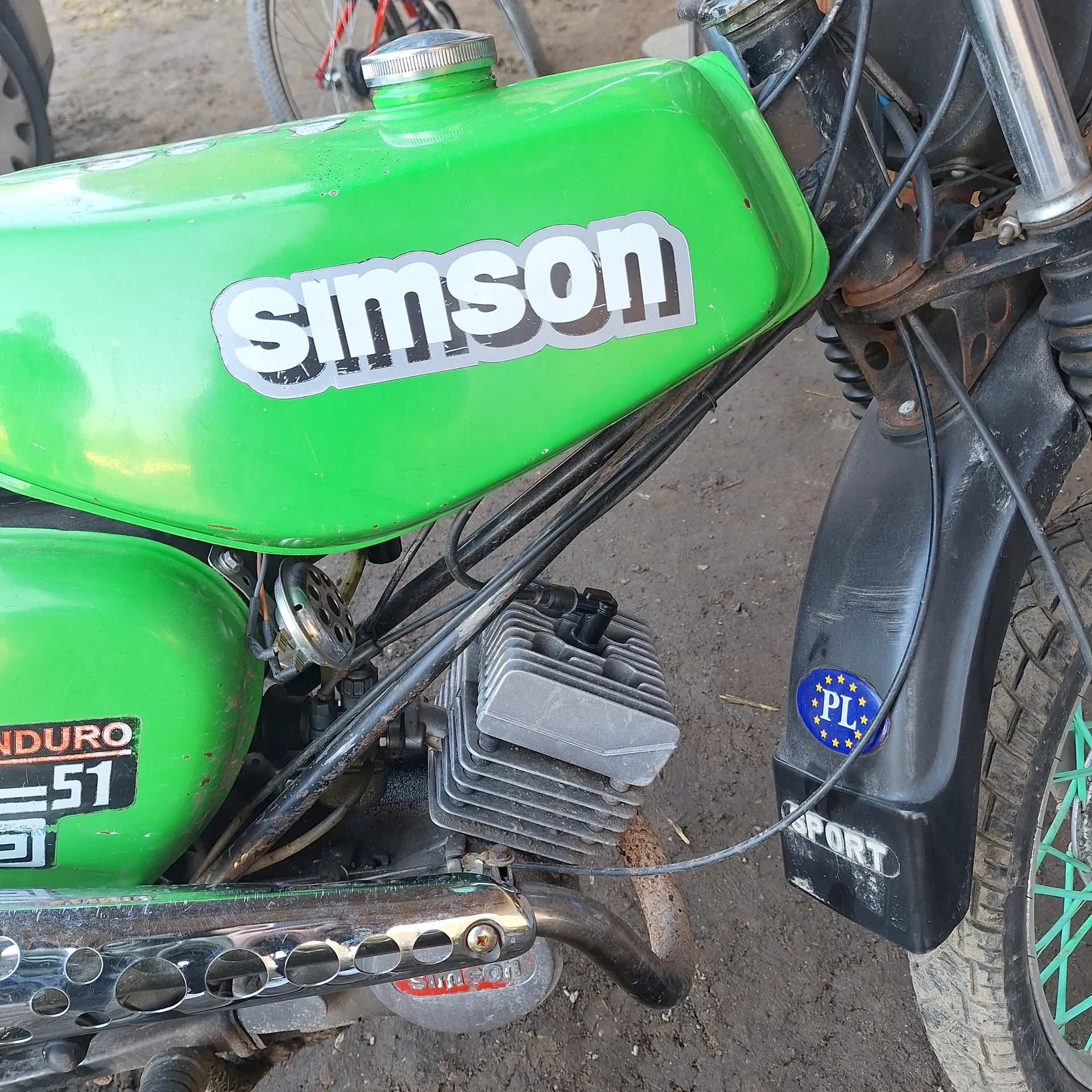 Zbliżenie na zielony motocykl marki Simson. Widoczny zielony zbiornik paliwa z białym napisem 'Simson', czarny element z napisem 'SPORT' oraz niebieską naklejką z literami 'PL' i żółtymi gwiazdkami. Obok znajduje się silnik, czarne przewody oraz zielony element z napisem 'DURO 51'. W tle fragment koła roweru z zielonymi promieniami i brudna ziemia.