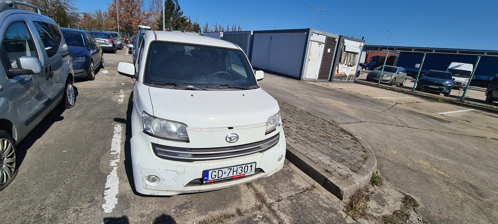 Biały samochód dostawczy z tablicą rejestracyjną GD-7H301 (PL) zaparkowany na asfaltowym parkingu. W tle inne pojazdy, przyczepy, kontenery przemysłowe oraz ogrodzenie z siatką. Niebo jest bezchmurne, a na ziemi widoczne są białe linie parkingu i fragmenty trawy.