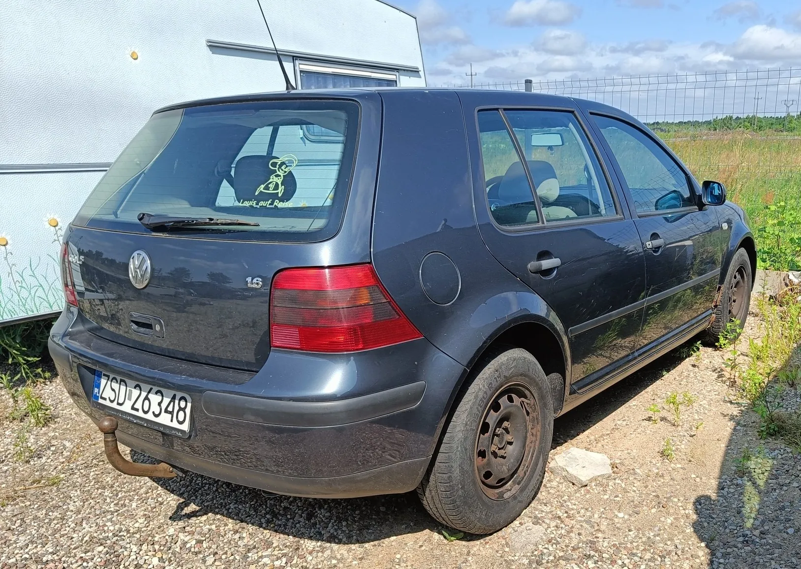 Ciemnoszary samochód marki Volkswagen Golf w wersji nadwozia hatchback, zaparkowany na żwirowym podłożu. Widoczny jest tył i prawy bok auta. Samochód posiada polską tablicę rejestracyjną o numerze ZSD 26348 oraz hak holowniczy. Na tylnej szybie znajduje się żółta naklejka z postacią psa i napisem „Let's roll”. W tle widoczny jest fragment białej przyczepy kempingowej oraz zielona roślinność pod jasnym, lekko zachmurzonym niebem.