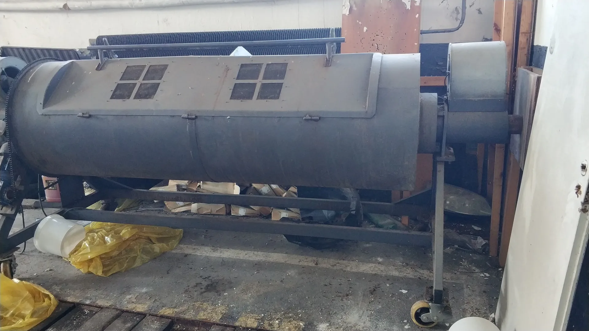 Duży, poziomy, metalowy cylinder przemysłowy z dwoma grupami czterech kwadratowych okienek na górnej części. Urządzenie zamontowane na metalowej ramie z kółkami. W tle betonowa podłoga, żółte worki plastikowe, pudełka kartonowe oraz elementy przemysłowe, w tym czarna rura i fragmenty ścian z widocznymi uszkodzeniami. Brak czytelnego tekstu.