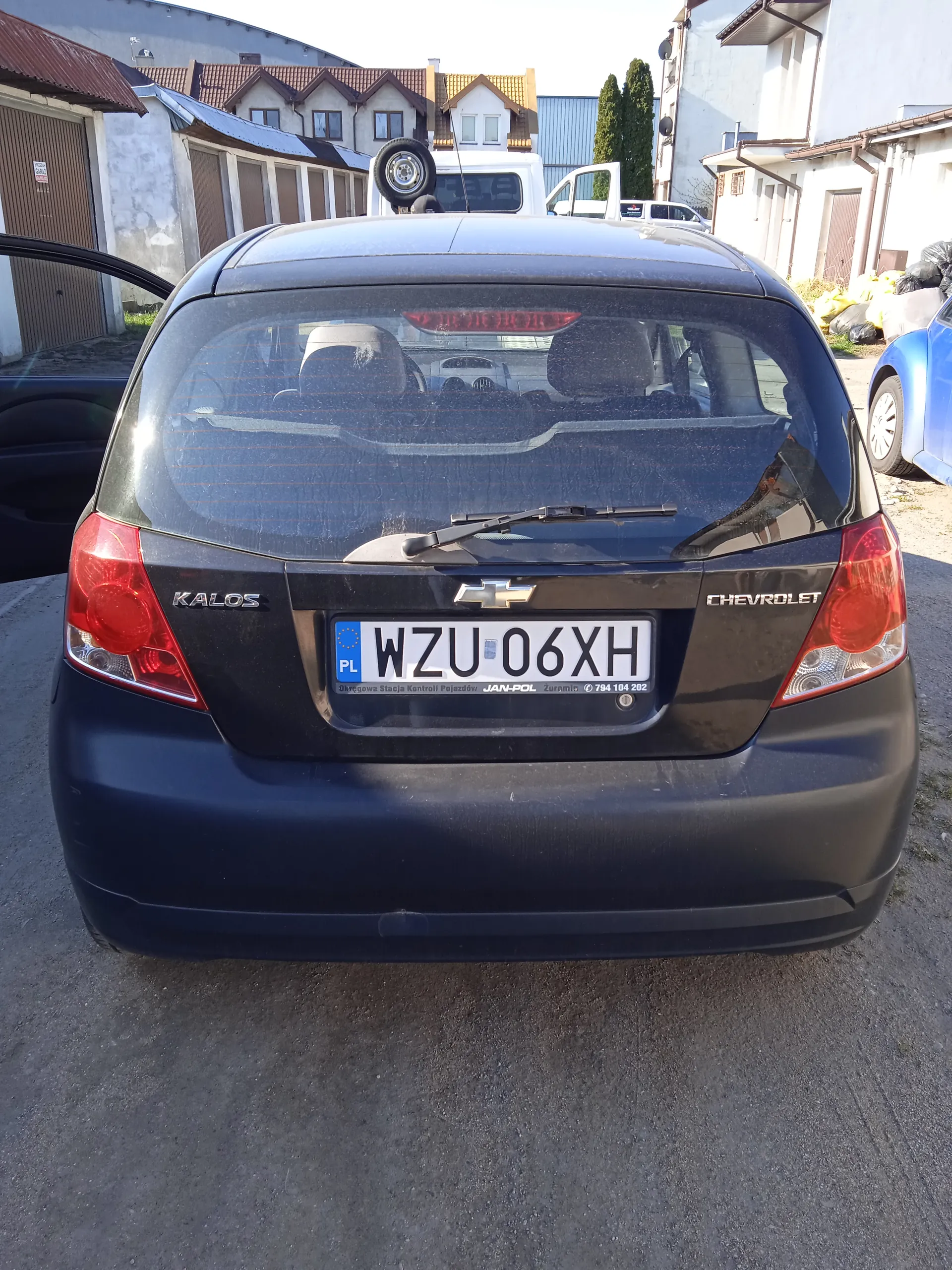 Z tyłu czarny samochód Chevrolet Kalos z tablicą rejestracyjną WZU 06XH. Po lewej stronie otwarte drzwi, na tle budynki mieszkalne i garaże, biały van z zapasowym kołem na dachu oraz niebieski samochód po prawej stronie. Tablica rejestracyjna zawiera napisy: PL, WZU 06XH, Ośrodek Szkolenia Kierowców JAN-POL, Zgierz, 794 104 202.