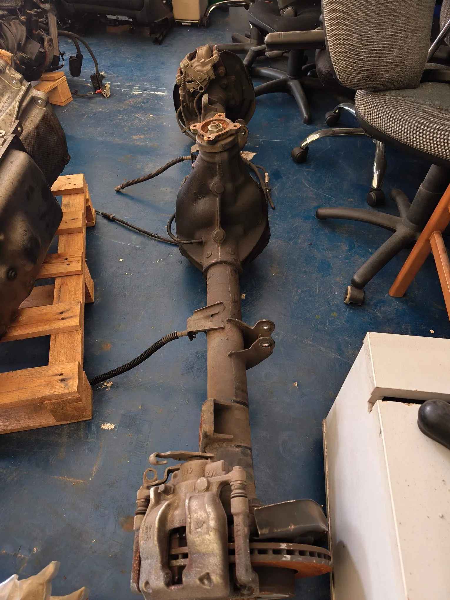 Zdjęcie przedstawiające metalowy element mechaniczny, prawdopodobnie oś samochodu z układem hamulcowym, leżący pionowo na niebieskiej podłodze w warsztacie. W tle widoczne są krzesła biurowe, drewniana paleta oraz inne części samochodowe.