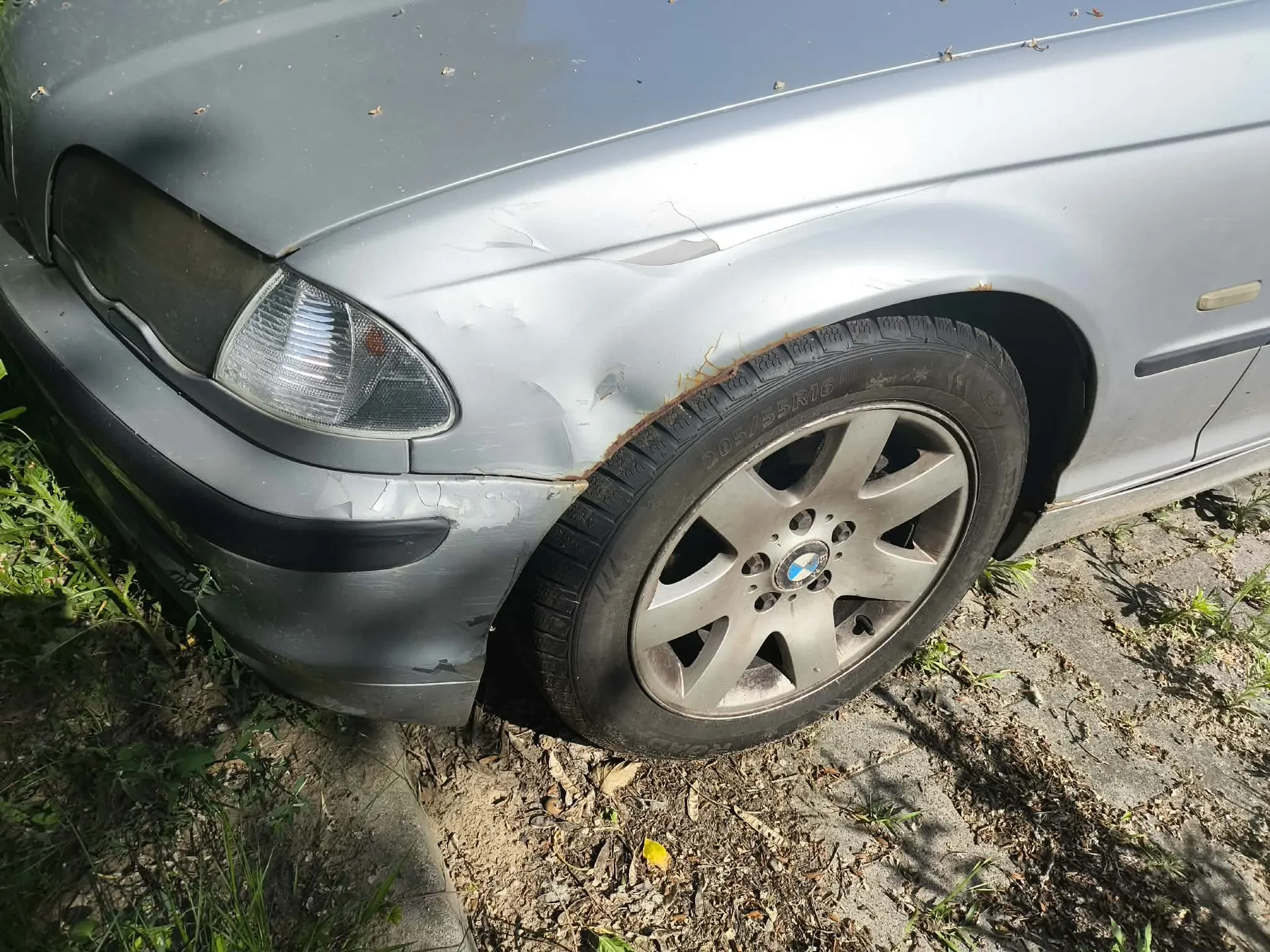 Zbliżenie na przedni lewy błotnik i koło srebrnego samochodu. Błotnik jest wgnieciony i porysowany nad nadkolem, widoczne są ślady korozji. Samochód posiada oryginalną felgę aluminiową z logo BMW w centrum. Pojazd stoi na nieutwardzonym podłożu z trawą i ziemią.