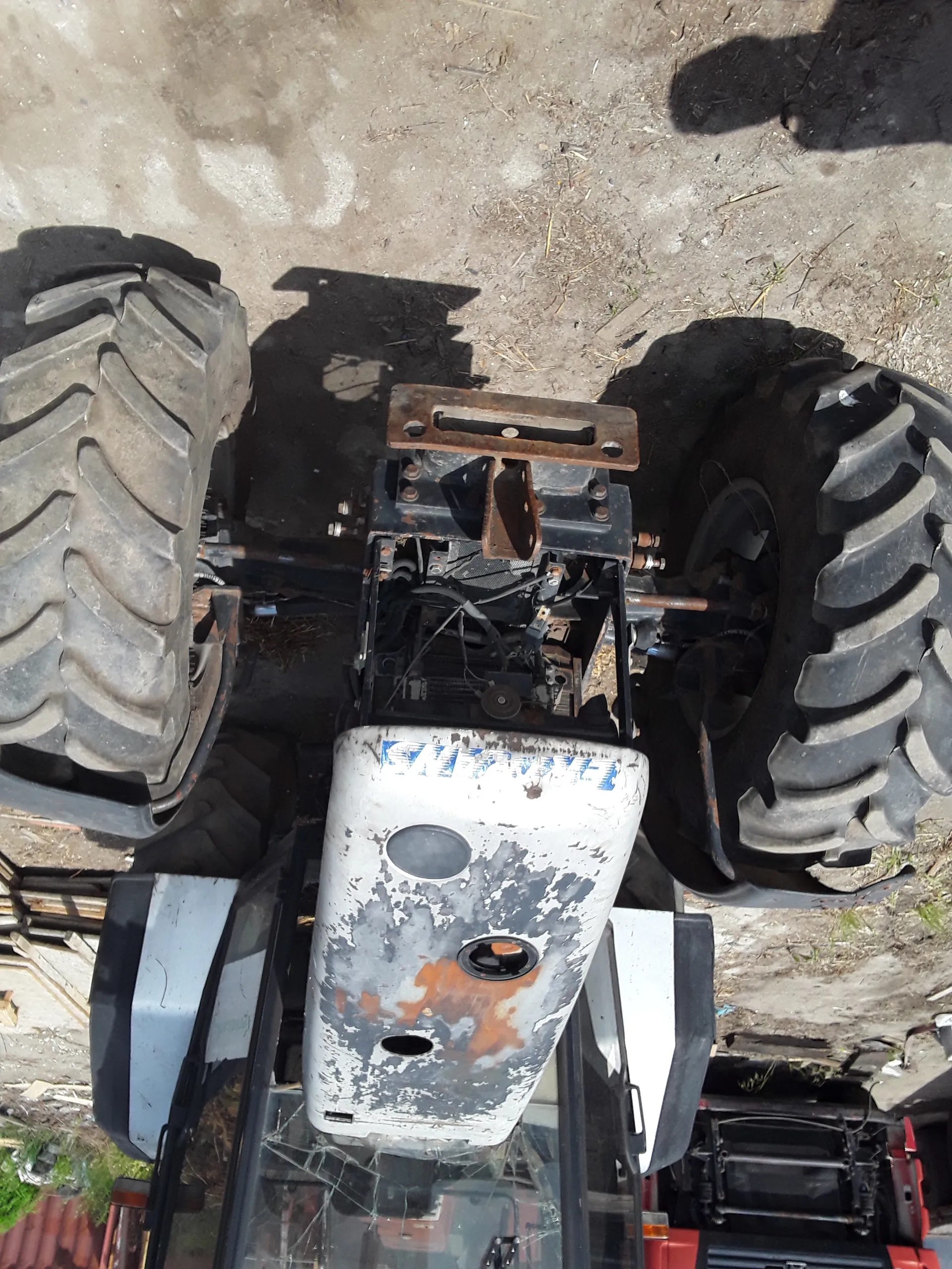 Zdjęcie traktora FENDT z widocznymi dużymi oponami o głębokich rynnach. Nadwozie traktora jest białe z miejscami rdzy i zarysowanej farby. Widoczne są części mechaniczne i przewody. W tle ziemia, czerwony pojazd oraz drewniane deski.