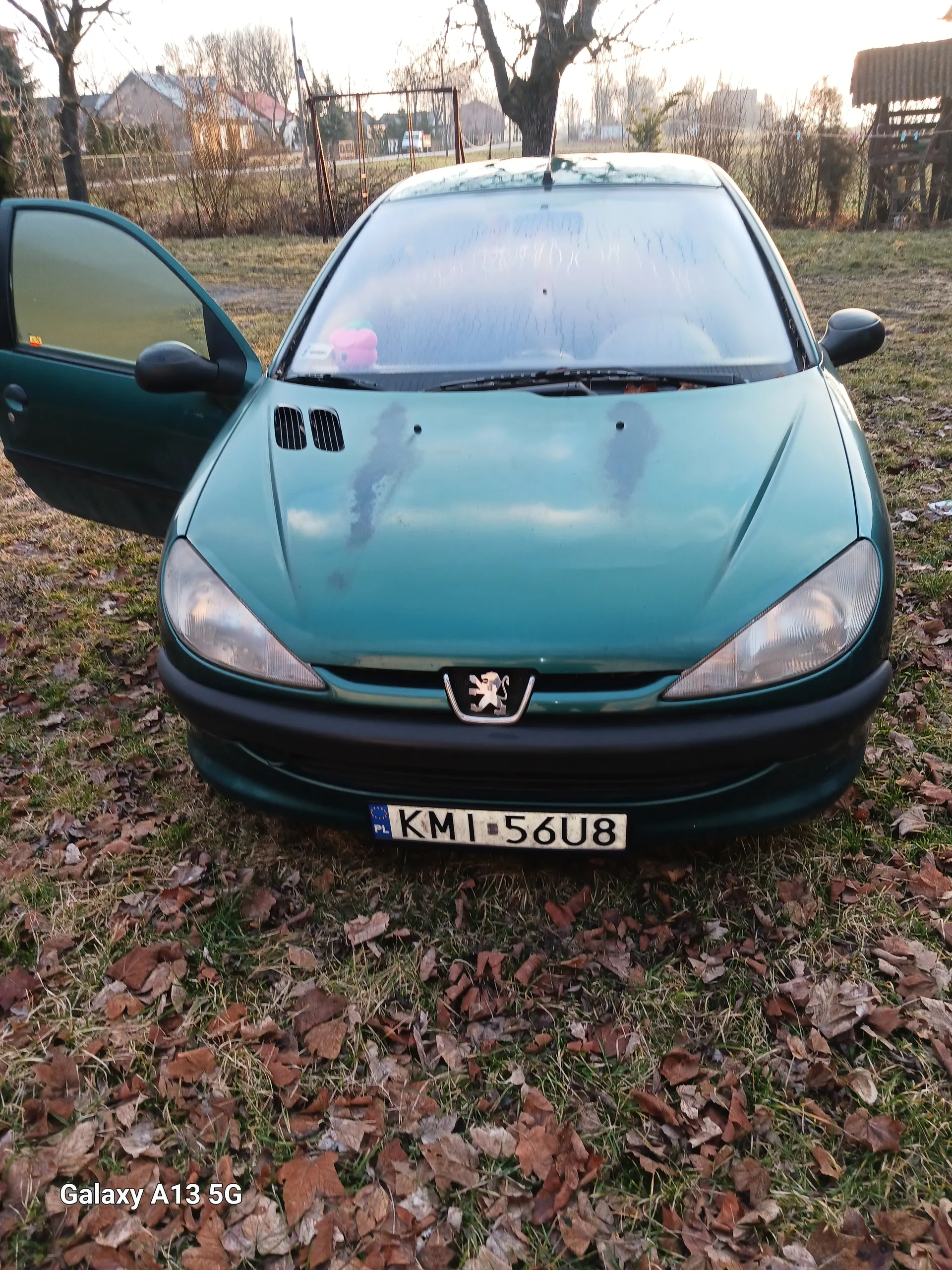 Zielony samochód marki Peugeot z otwartą lewą przodem drzwią, zaparkowany na trawie pokrytej suchymi liśćmi. Widoczna tablica rejestracyjna KMI 56U8 oraz logo marki na masce. W tle drzewa, budynki i ogrodzenie. W prawym dolnym rogu obrazu widoczny napis Galaxy A13 5G.