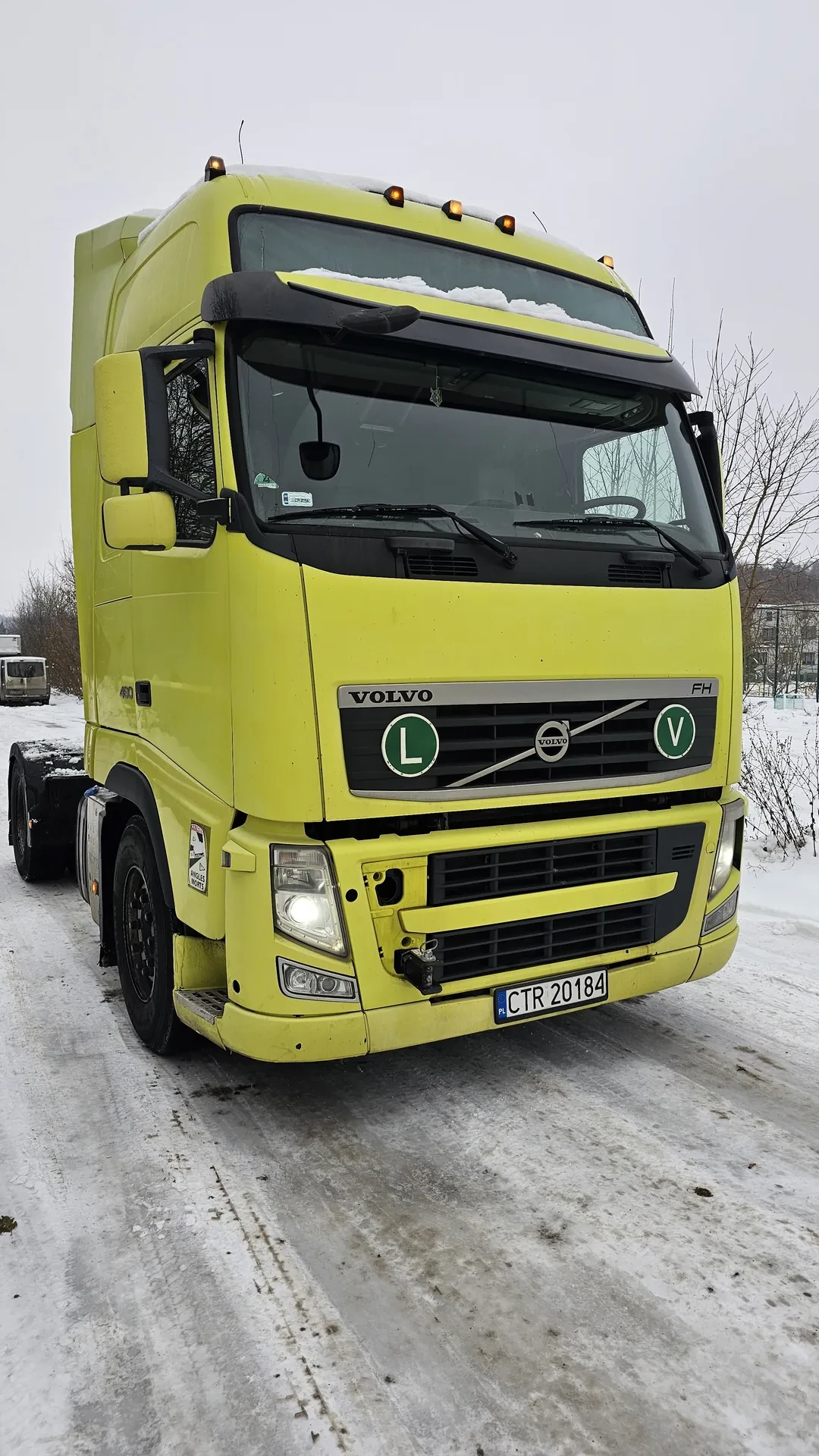 Żółty ciężarówka Volvo FH z tablicą rejestracyjną CTR 20184 (PL), zielonymi tabliczkami "L" i napisem "FH" na masce. Zaparkowana na śnieżnej drodze z odciskami opon. Widoczne włączone reflektory, lusterka, śnieg na przedniej szybie. Tło: nagie drzewa, budynki.