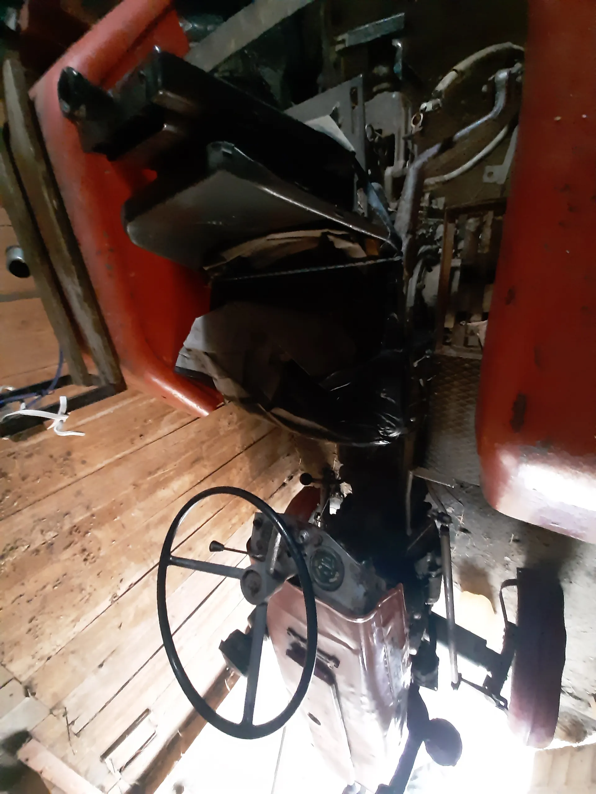 Stary traktor z czarną kierownicą, czerwonymi metalowymi częściami, czarną torbą na siedzeniu oraz widocznymi elementami mechanicznymi. Tło stanowi drewniana ściana z pionowych desek. Wnętrze przypomina stodołę lub garaż.