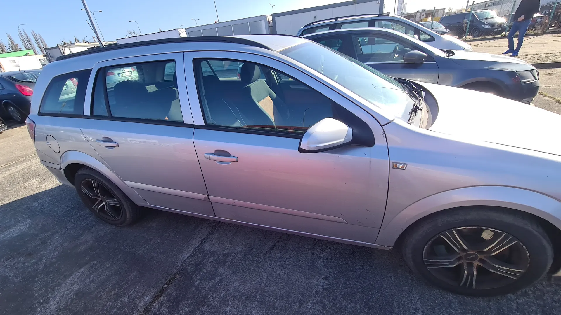 Srebrny samochód typu kombi zaparkowany na asfaltowym placu. W tle widoczne są inne pojazdy, osoba stojąca w odległości, budynki oraz słoneczne niebo. Samochód ma czarne felgi i ciemne szyby. Na tle znajdują się również latarnie uliczne i ogrodzenie.